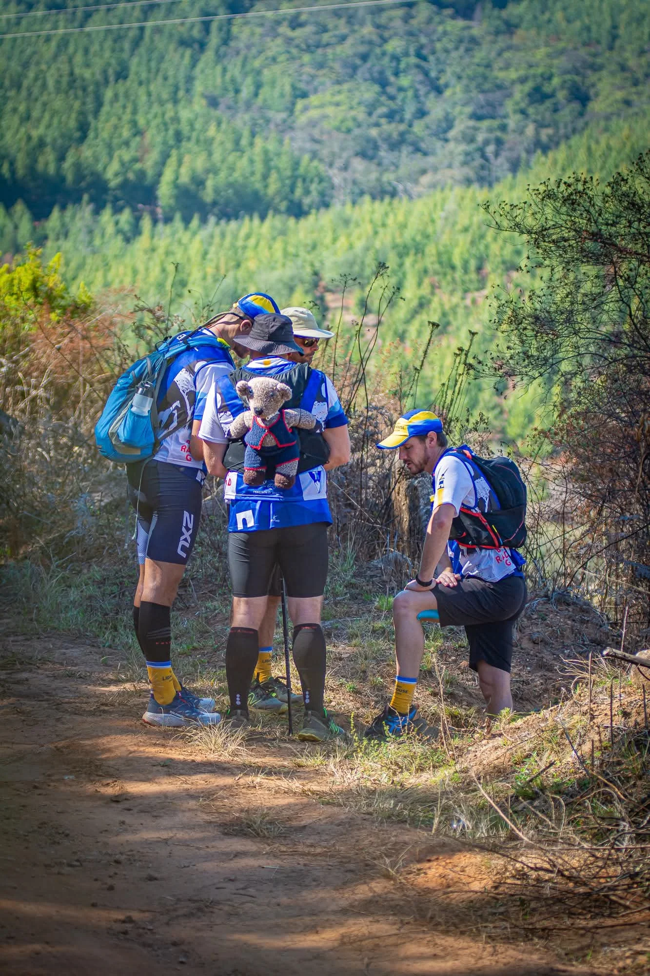 Four men in athletic gear, some with hats, are on a hiking trail in a forested area, with one of them holding a teddy bear dressed in clothing.