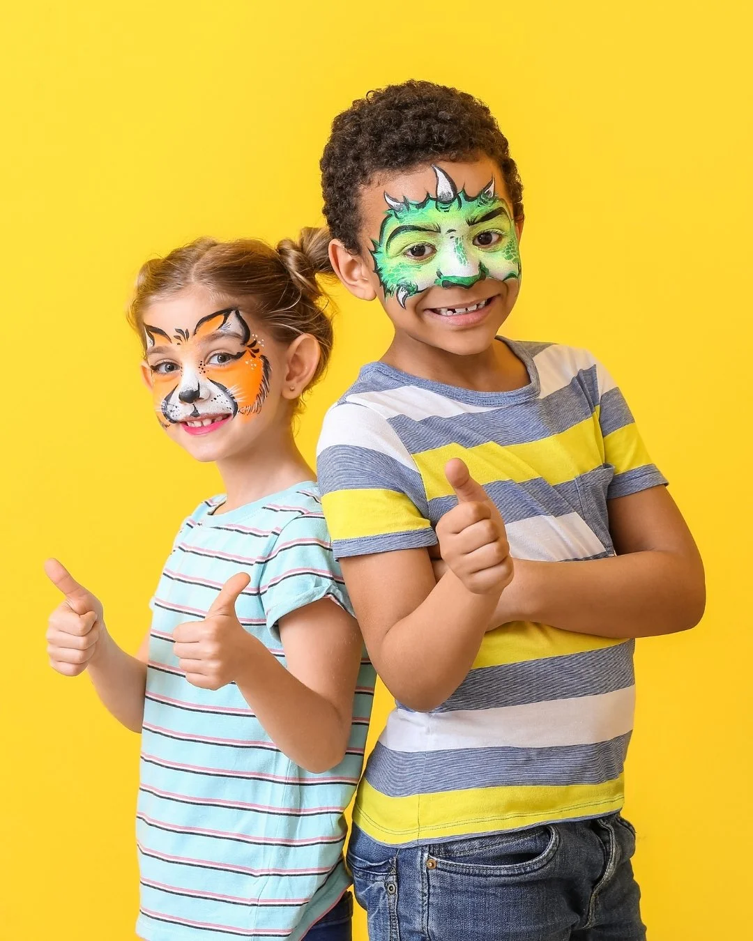 Two children with face paint, smiling and giving thumbs up, standing against a yellow background.