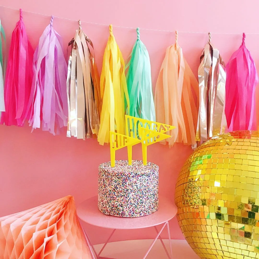 Colorful birthday party decorations including pastel tassel banners on a pink wall, a birthday cake with multicolored sprinkles and a yellow 'Happy Birthday' banner on top, a large gold disco ball, and pastel-colored honeycomb paper decorations.