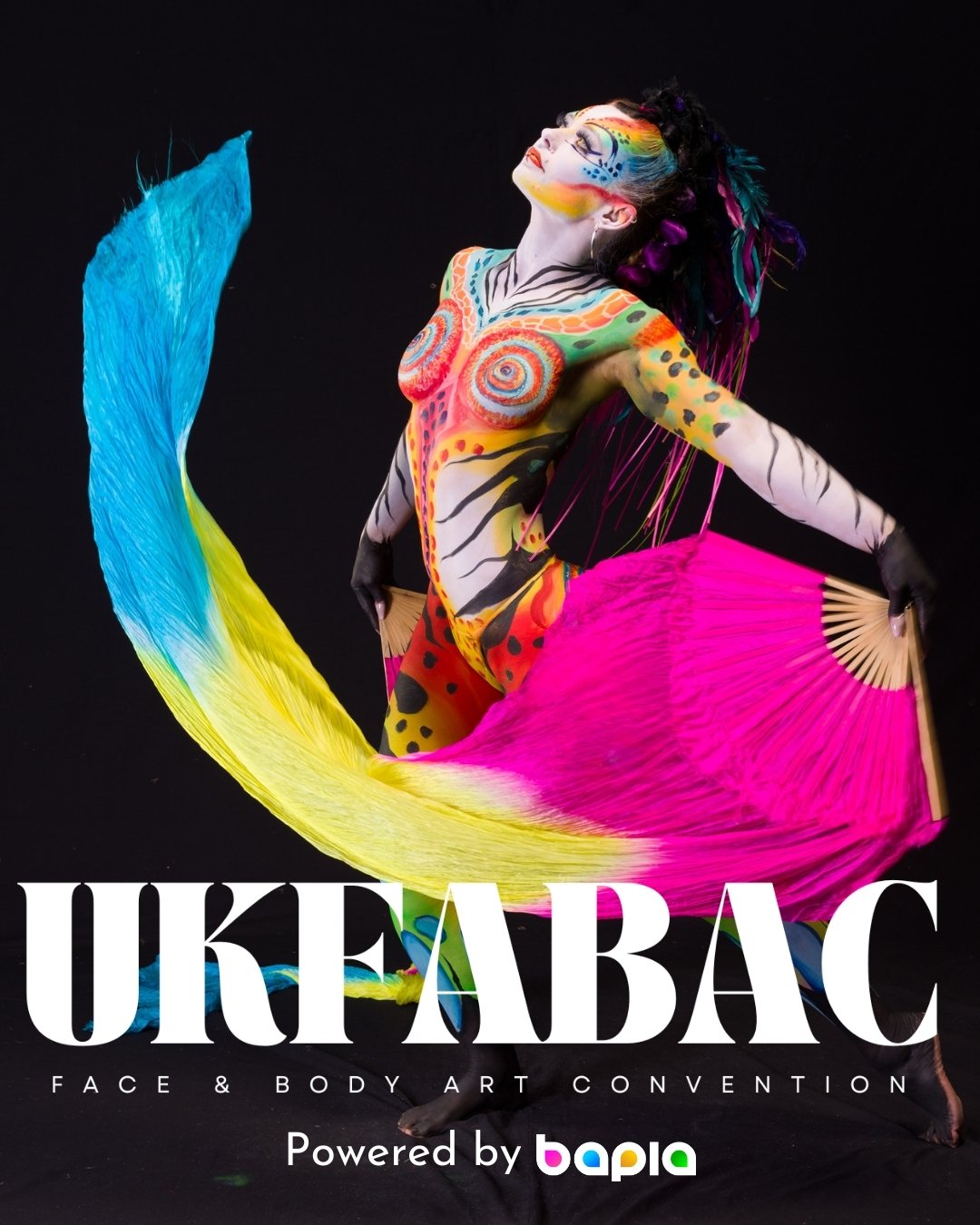 A performer in colorful body paint and costume dancing with large pink and blue fans at an event called UKFABAC, Face & Body Art Convention.