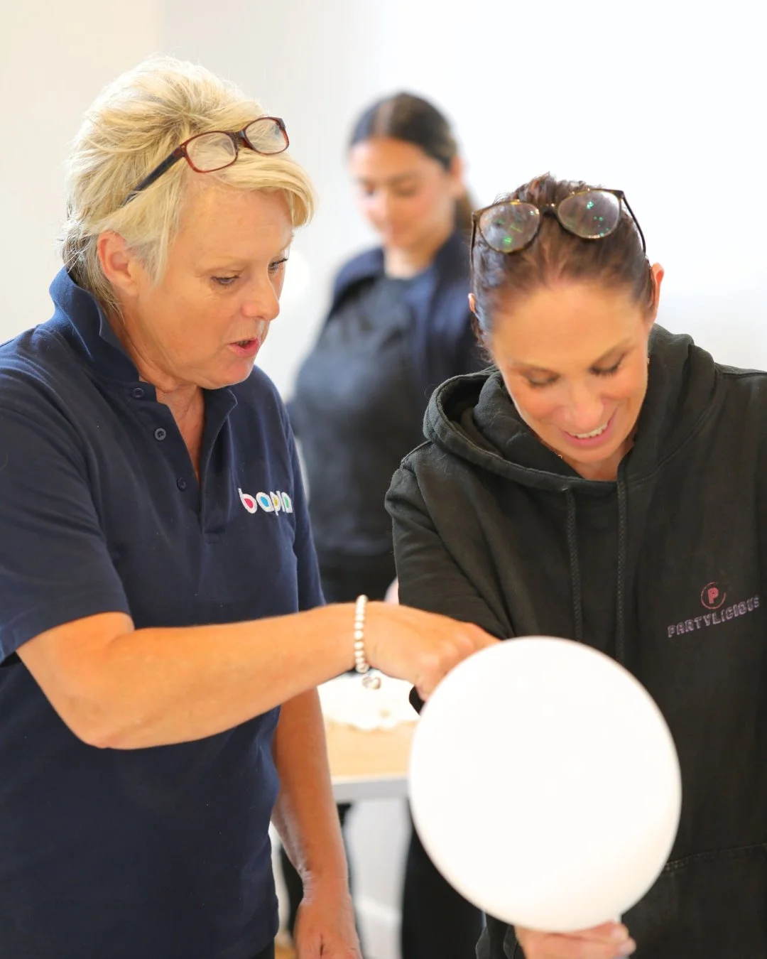 Three women at a social event, with two in focus. The woman on the left has short blonde hair, glasses on her head, and is wearing a navy shirt, engaging with the woman on the right who has dark hair tied back, glasses on her head, and is wearing a black hoodie. They appear to be talking, and the woman on the right is holding a white balloon. A third woman in the background is slightly blurred.