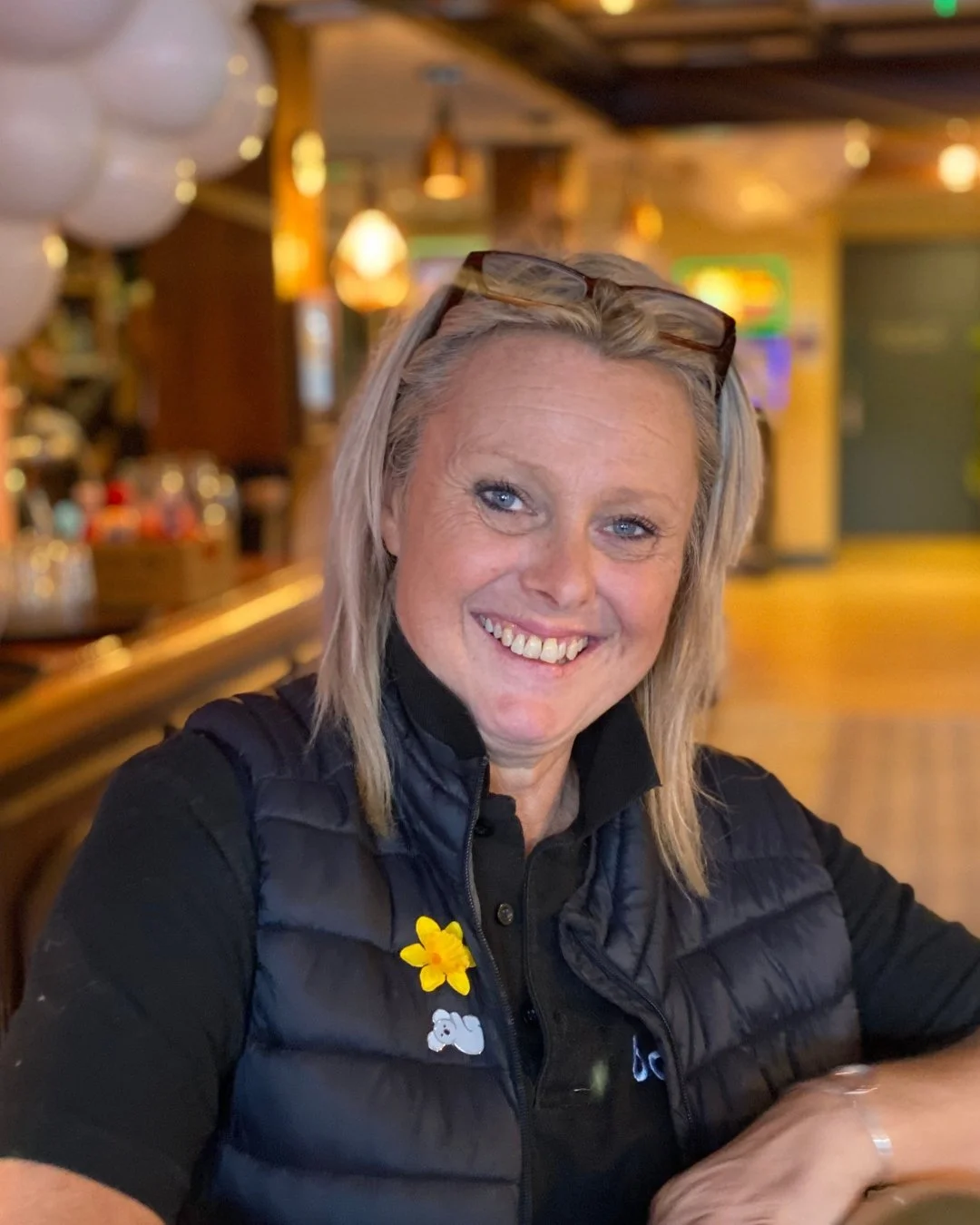 Smiling woman with blonde hair wearing glasses on her head and a black vest, sitting inside a warmly lit bar or restaurant