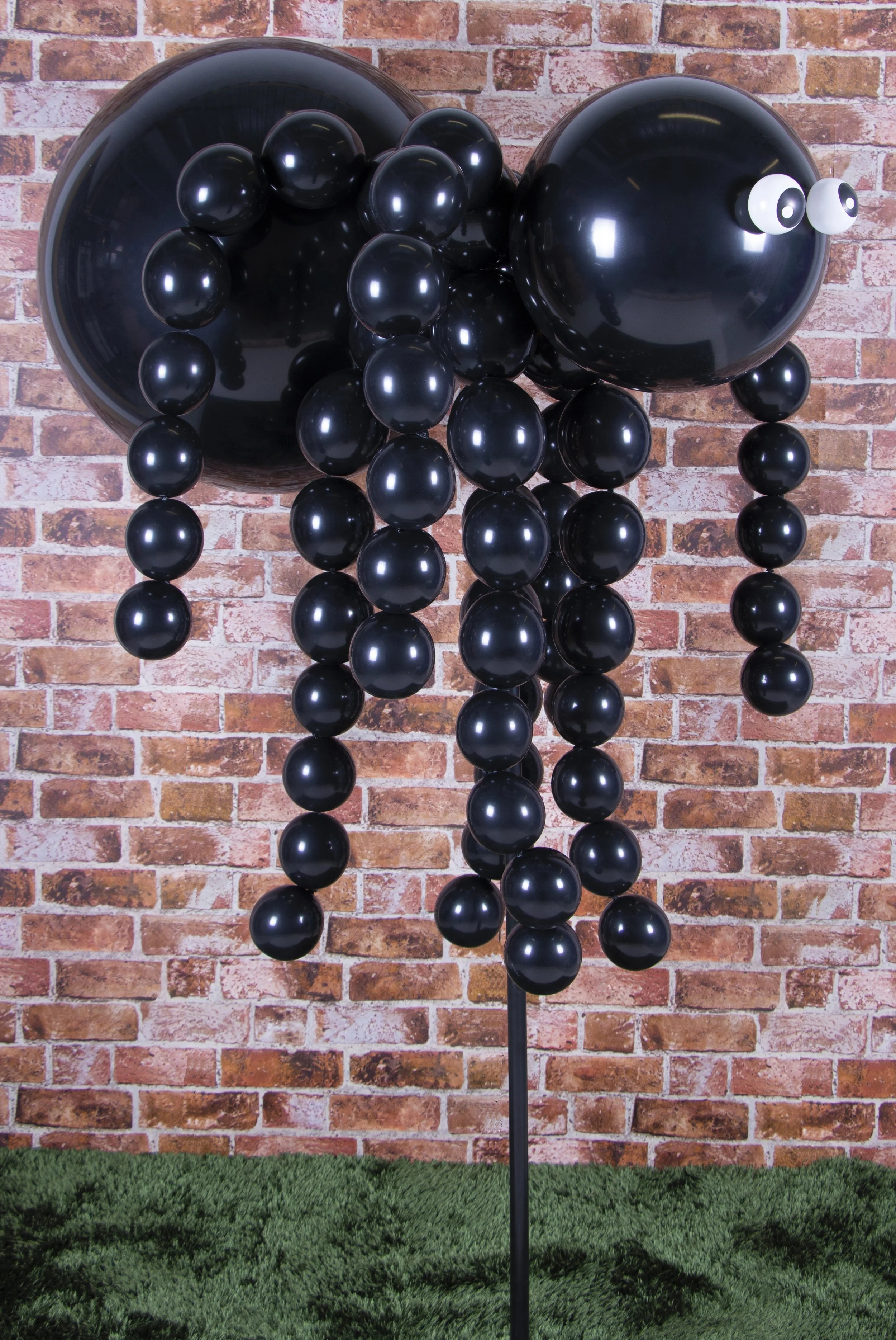 Balloon sculpture of a spider with a black spherical body, a large head, long hanging legs, and googly eyes, set against a brick wall background.