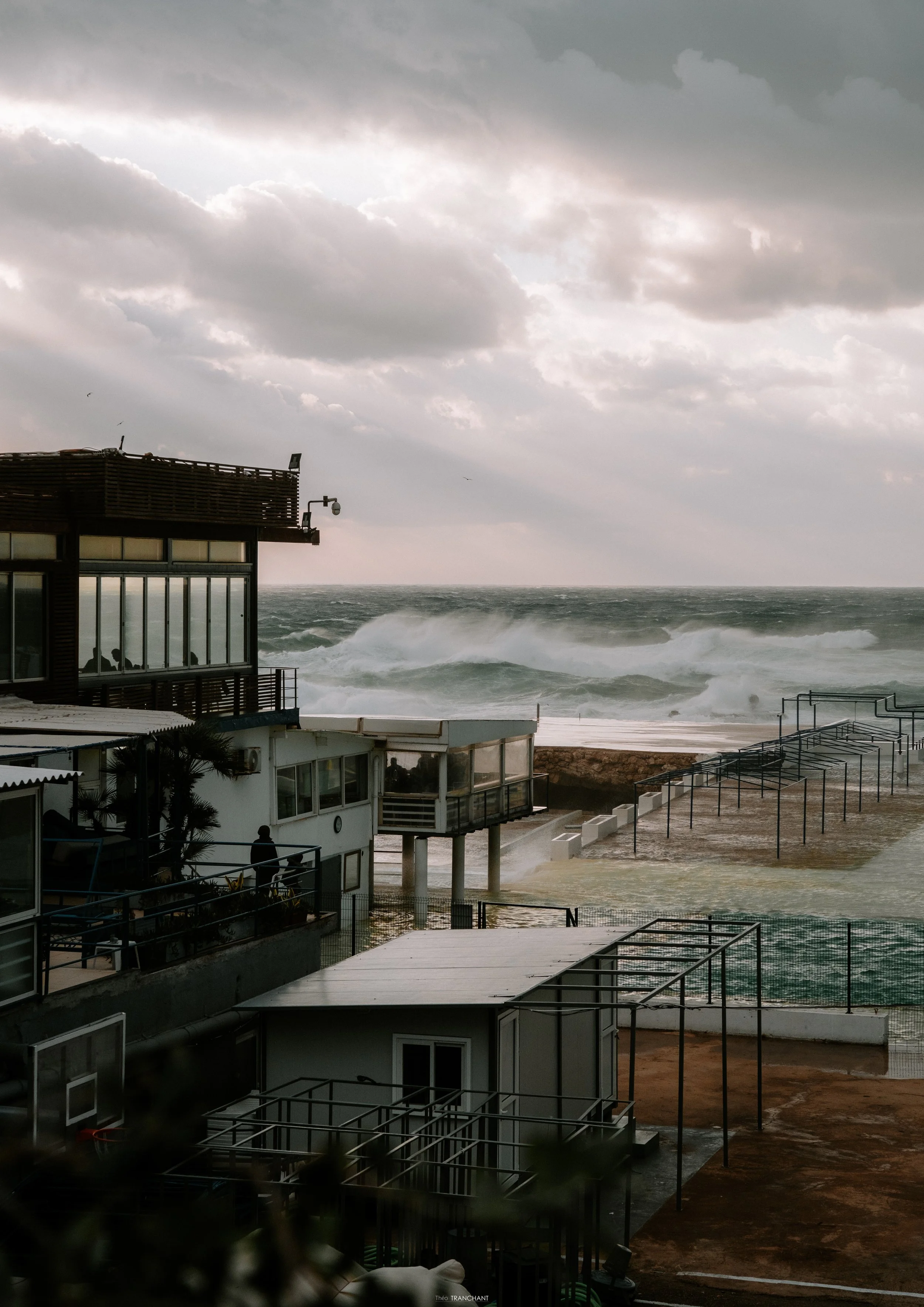 Overcast sky with dark clouds over a stormy ocean, with a building and a promenade on the shore, while large waves crash against the coast.