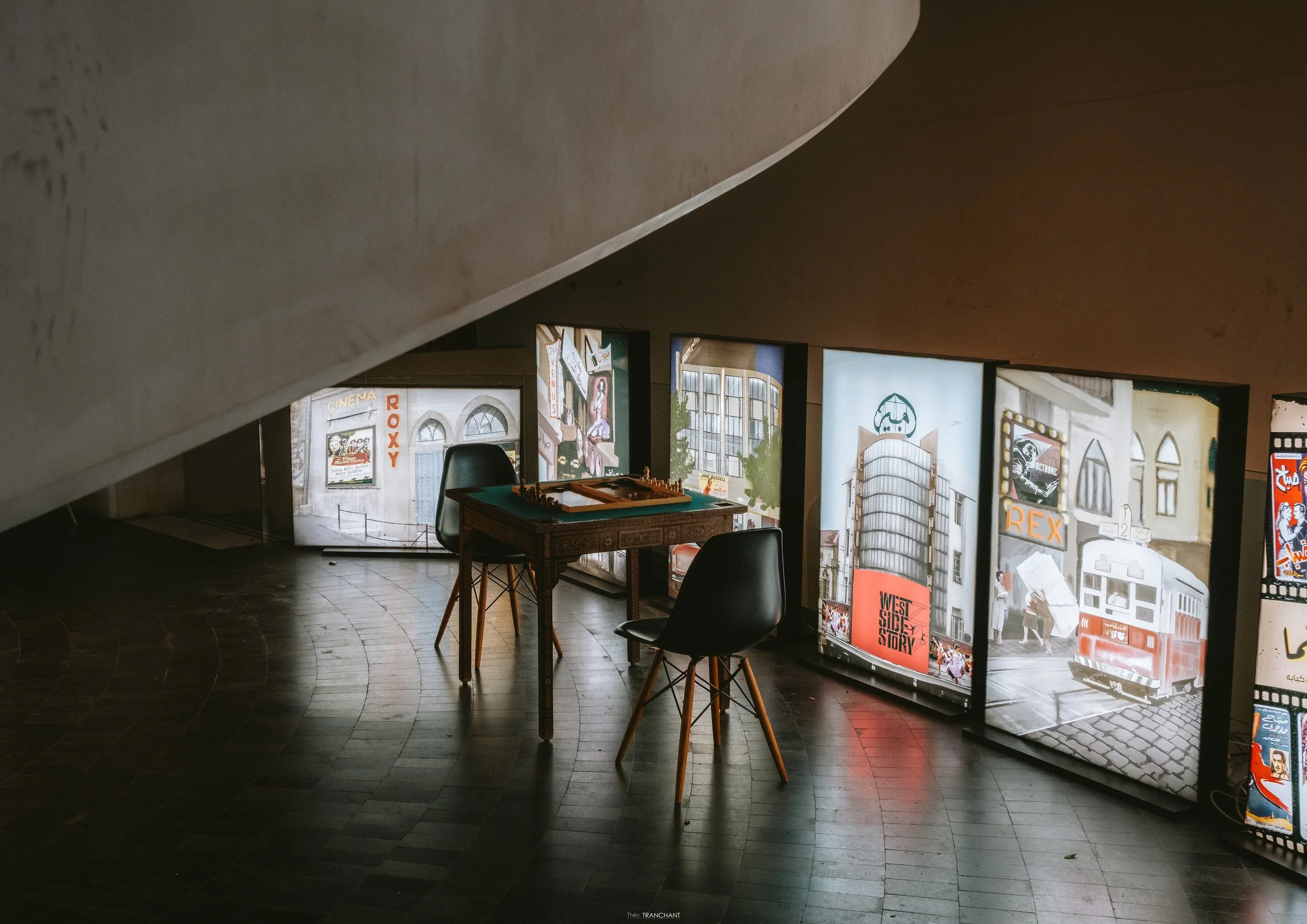 Indoor scene of a room with a sloped ceiling and a row of illuminated posters or screens displaying various images and text, including a chessboard on a table with chairs.