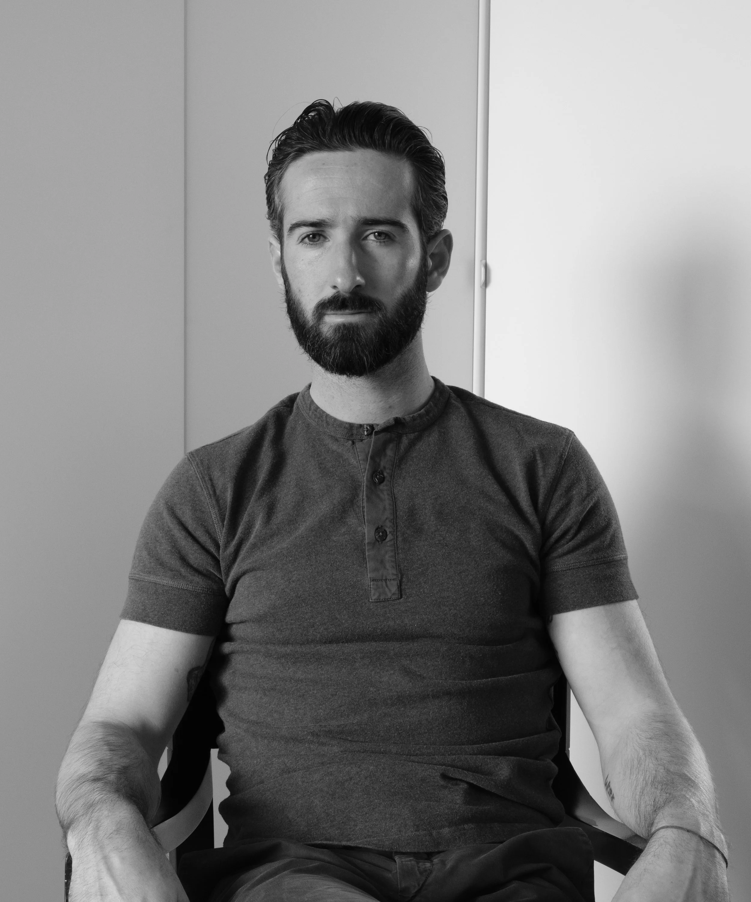 Black and white photo of a man with slicked-back hair and a beard, sitting in front of a plain background, wearing a dark collared shirt with a buttoned placket.