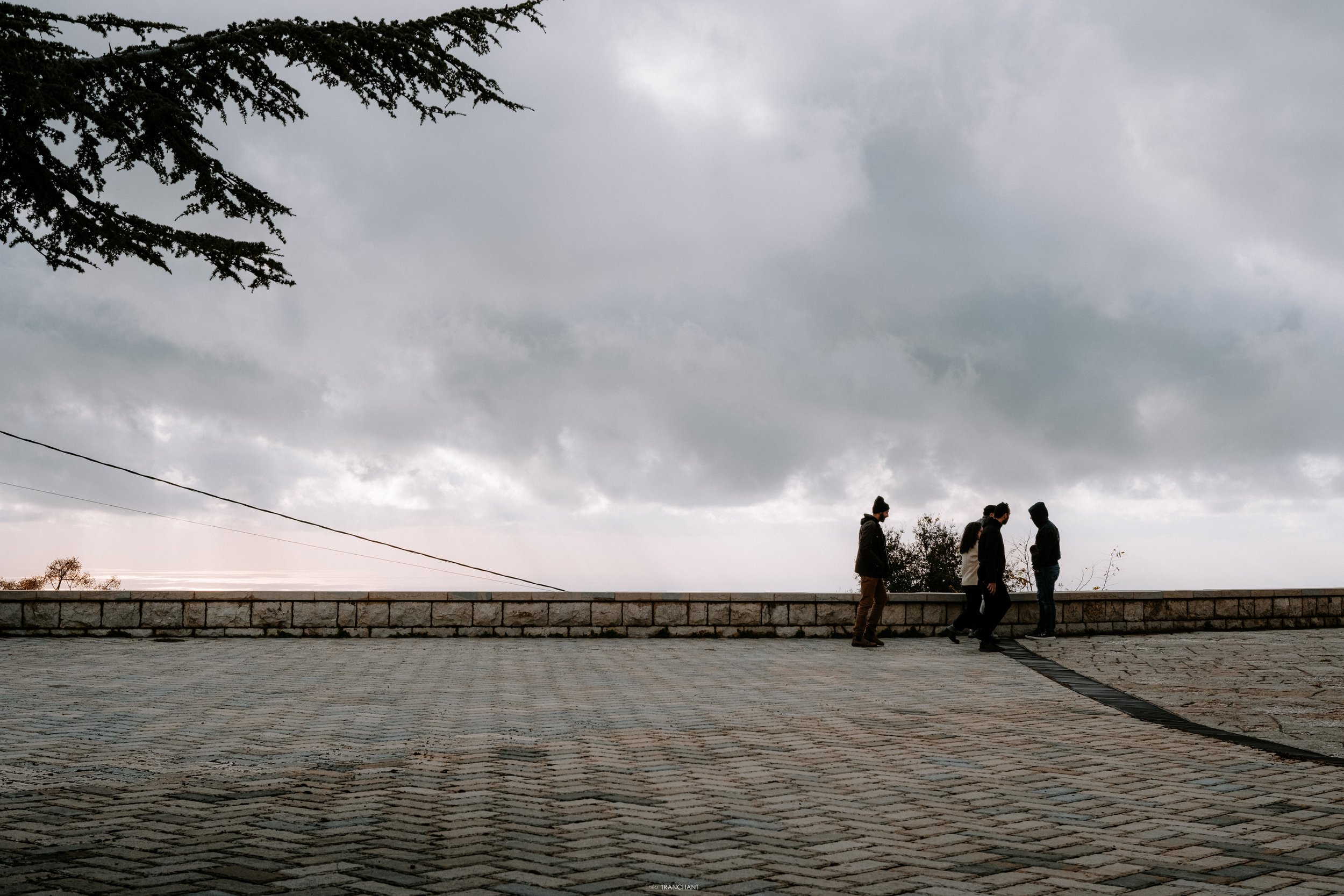 Overcast sky with dark clouds above a brick wall and a group of five people standing on the pavement, some walking.