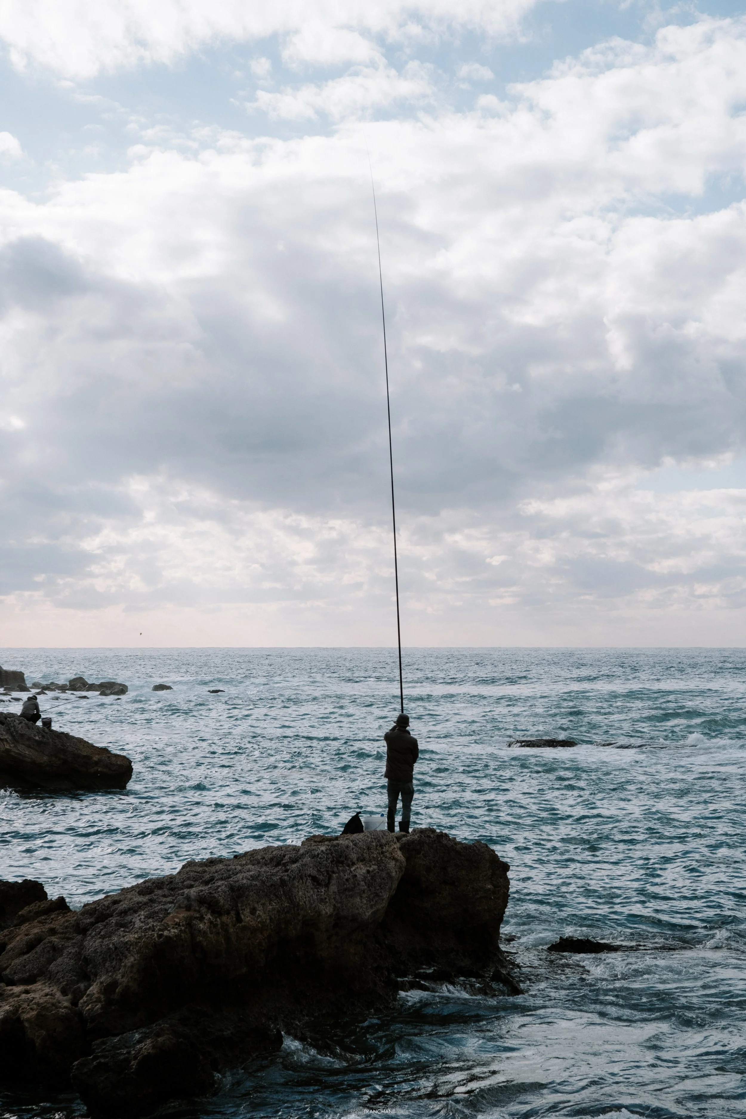 A person fishing off rocks by the ocean on a cloudy day, with a line extending into the water.