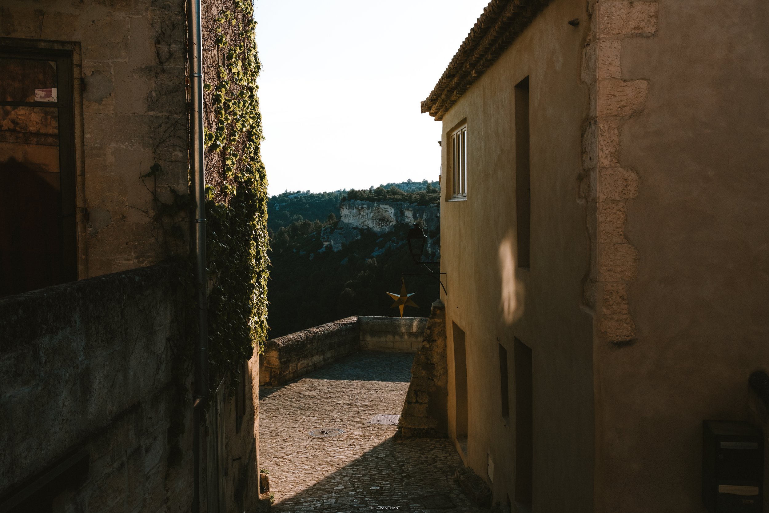 A narrow cobblestone alleyway between old stone and stucco buildings with a view of a distant hillside with white cliffs and green trees in the background.