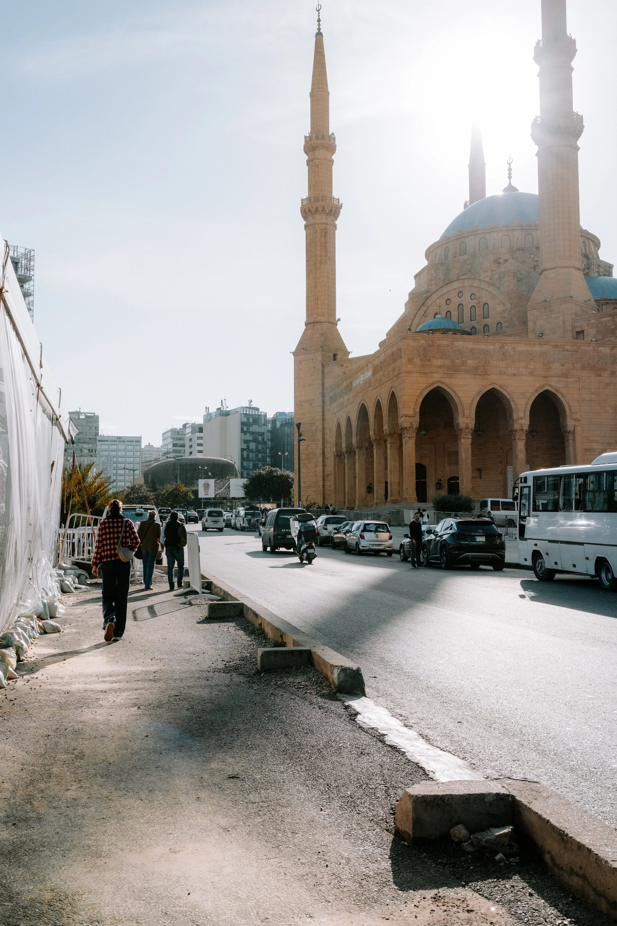 People walking along a sidewalk in front of a mosque with blue domes and tall minarets, with cars parked on the street and modern buildings in the background.
