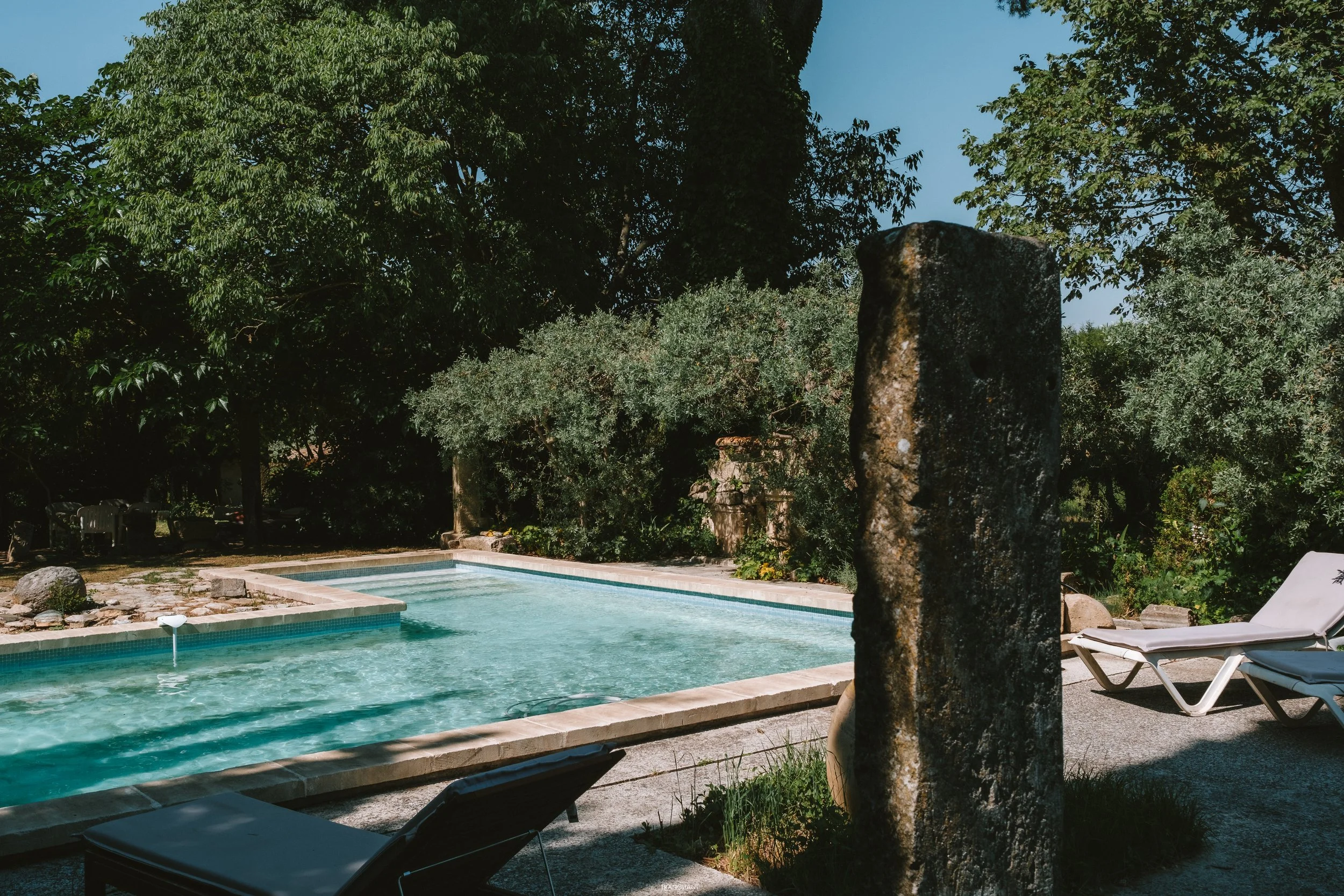 View of a backyard swimming pool with lounge chairs, lush green trees, and a large stone feature.