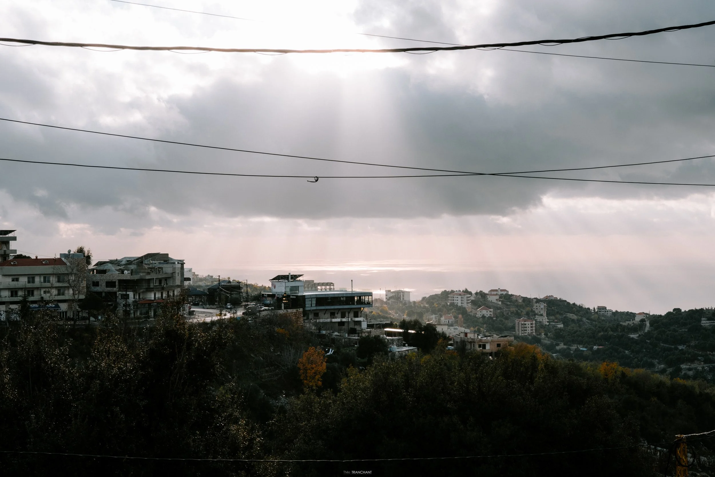 Overcast sky with dark clouds over a cityscape and hilly terrain, power lines in the foreground, and buildings below.