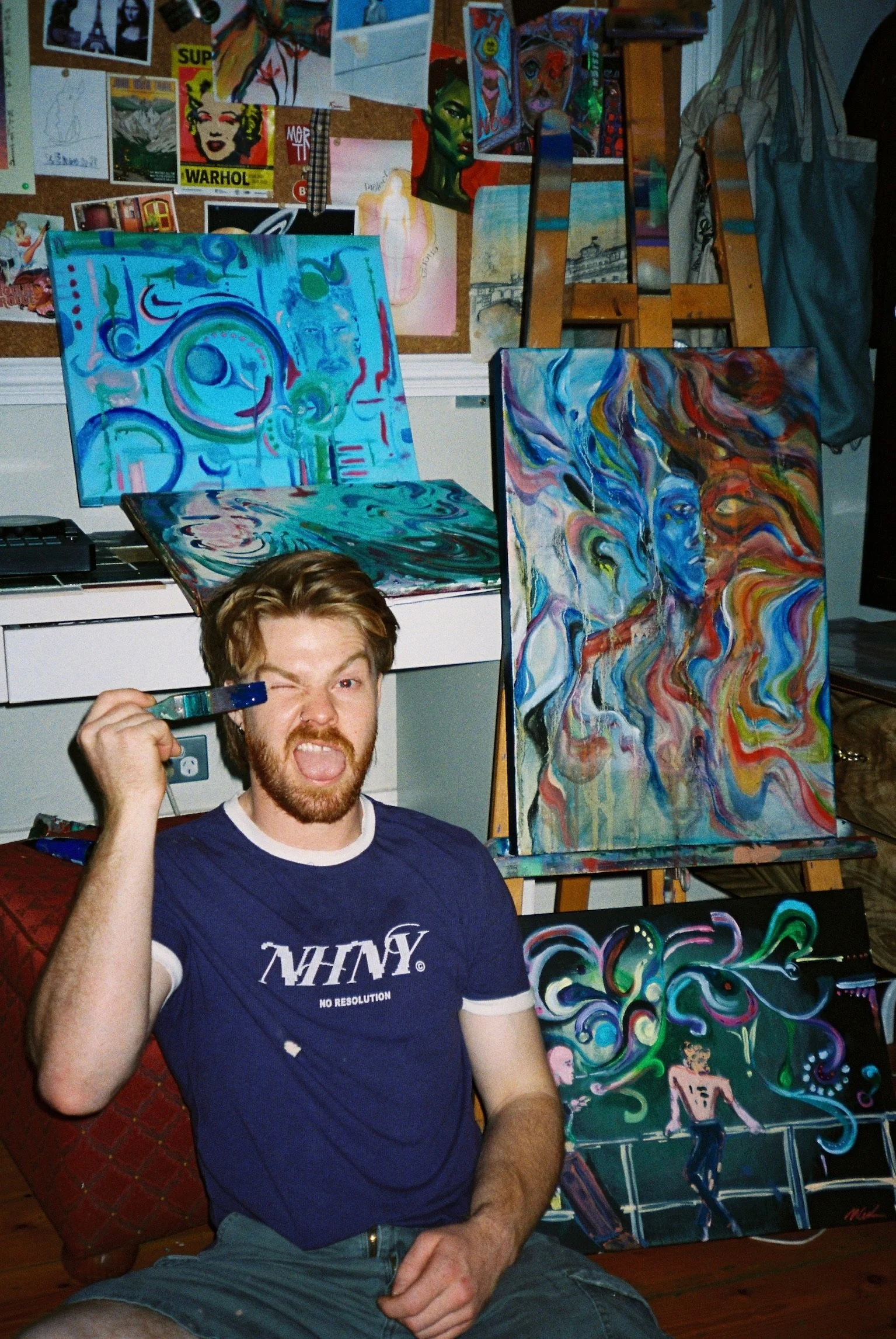 A young man with a beard and brown hair, wearing a navy blue T-shirt and gray shorts, is sitting on a red patterned chair in front of a table. He is holding a paintbrush in his right hand, which is near his face, and has a playful expression with one eye closed and tongue sticking out. Behind him, there are several colorful abstract paintings on easels, featuring swirling patterns and vibrant colors. The background includes a wall with pinboards full of various artwork, posters, and photographs.