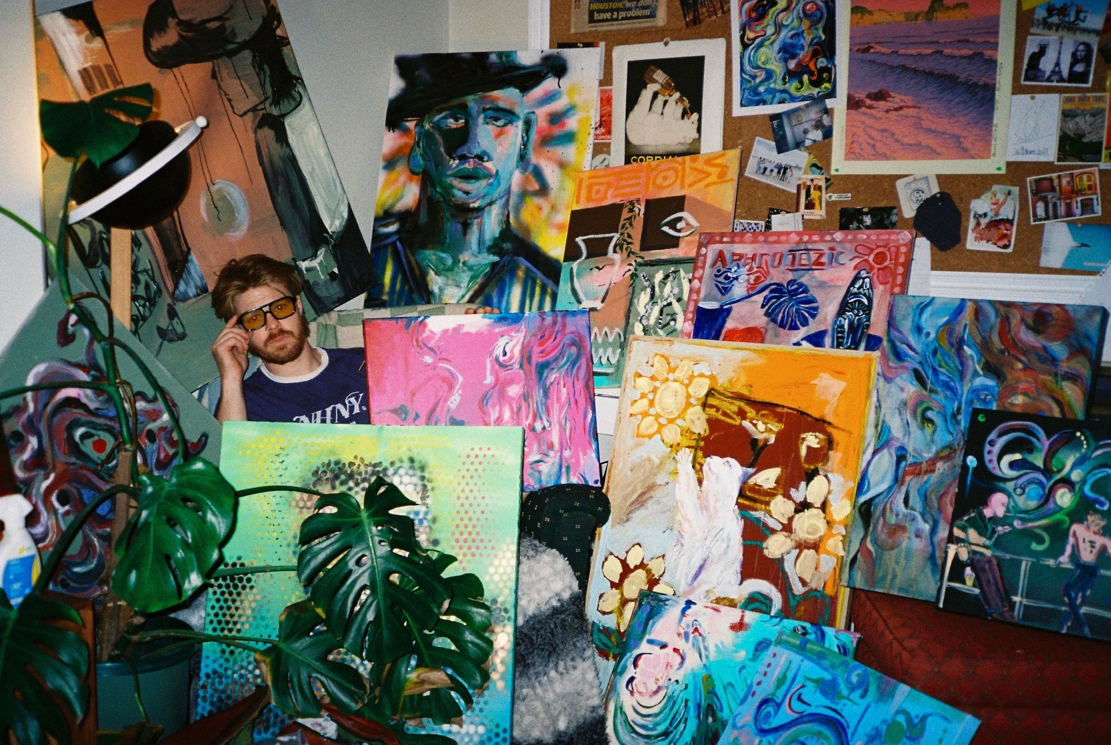 An art studio with multiple colorful paintings leaning against walls and displayed on tables. A man with glasses and a beard, wearing a T-shirt with the word 'WHY,' is seated in the foreground. The background features a bulletin board with various photos and artwork, and a large leafy plant partially obscures some of the artwork.