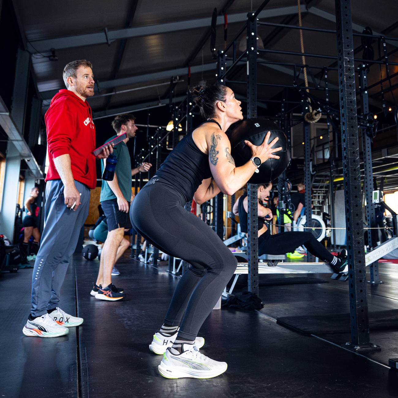 Vrouw doet squats met medicijnbal tijdens groepsfitnessles in sportschool, omringd door mensen die achter haar oefeningen uitvoeren.