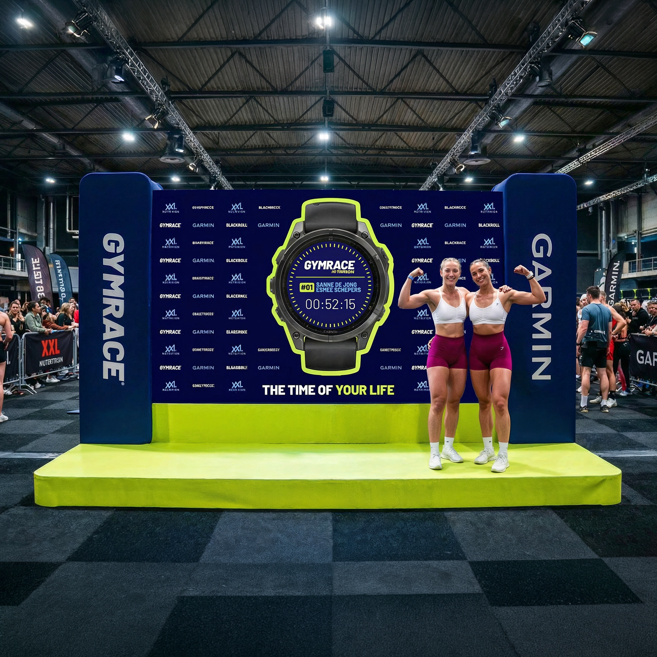 Twee vrouwen poseren met gespierde armen in sportoutfit, in een sportevenement met een grote Garmin-Gymrace-banner en een groot horloge-afbeelding op de achtergrond.