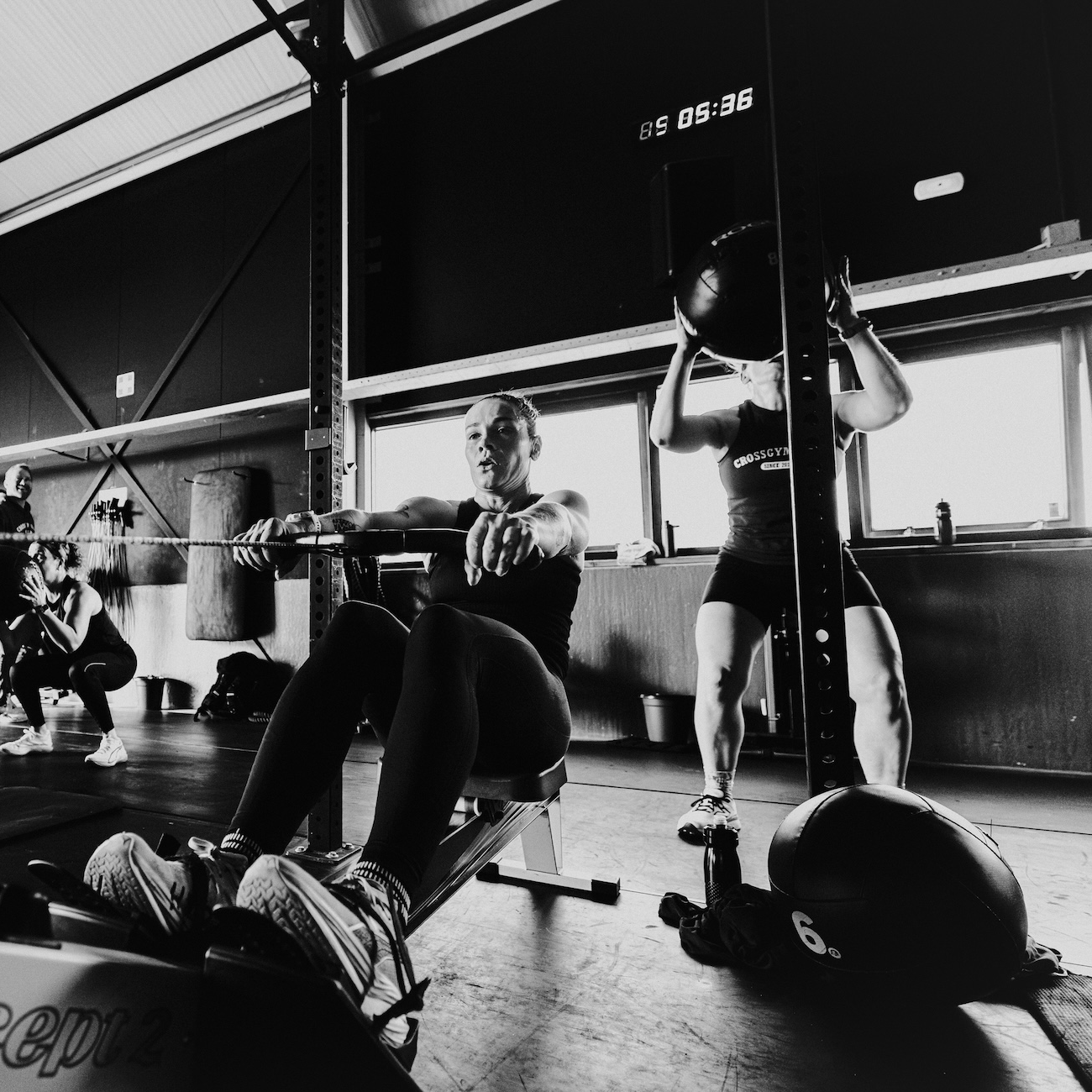 Mensen trainen in een gym, een vrouw die met een kabeltrekker oefent en een vrouw die met een zandzakje op haar schouders squat.