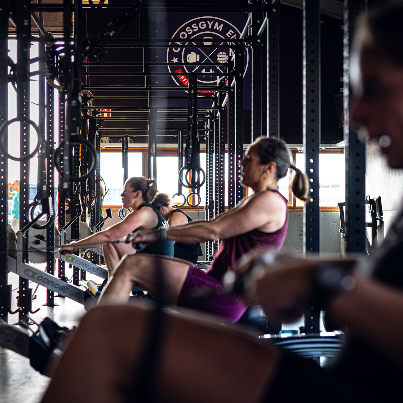 Mensen die in een sportschool op roeimachines trainen, met het logo 'CrossGym' op de achtergrond.