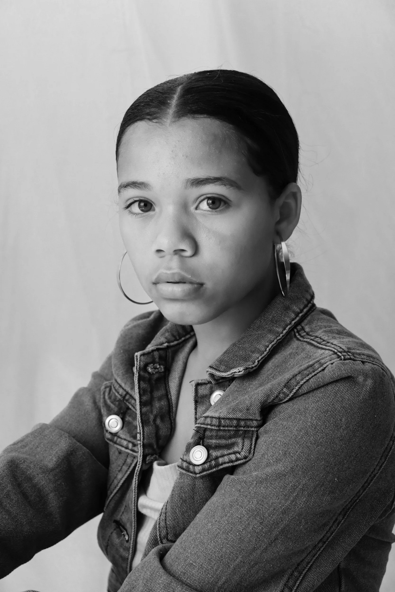 A young woman with dark hair styled in a sleek, middle-parted bun, wearing large hoop earrings, a denim jacket over a light-colored top, and strikingly expressive eyes, seated against a plain light background.