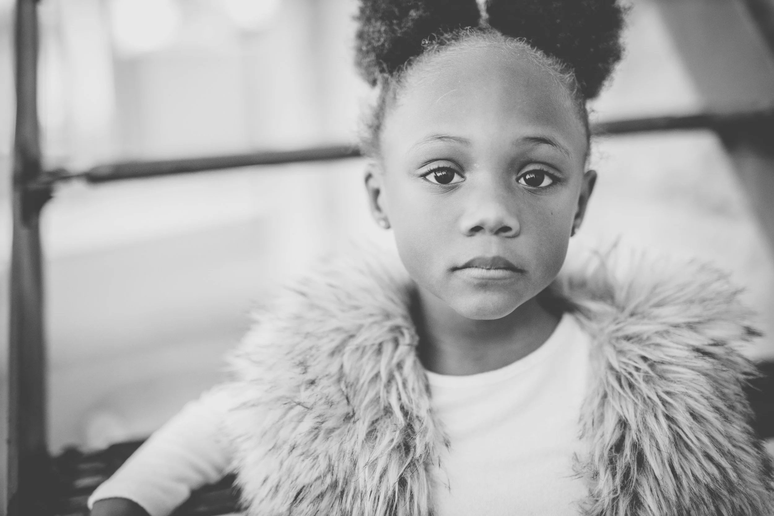 Close-up of a young girl with curly hair and expressive eyes, wearing a fluffy collar, looking directly at the camera, in black and white.