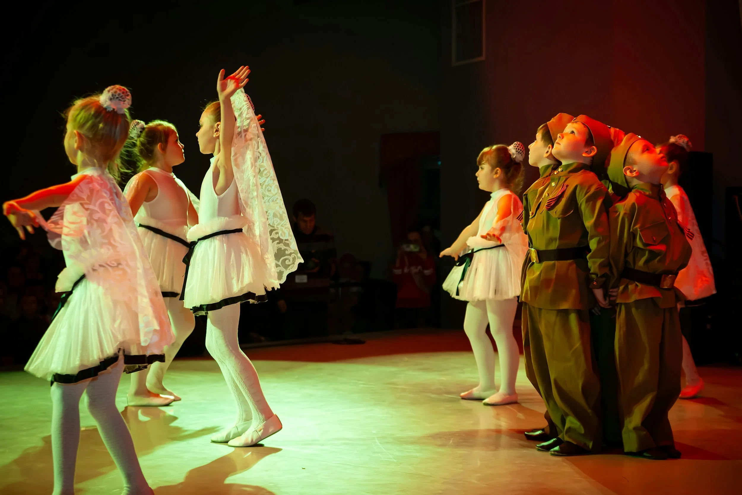 Children dressed in ballet costumes and military uniforms performing on stage, with spotlights and a dark background.