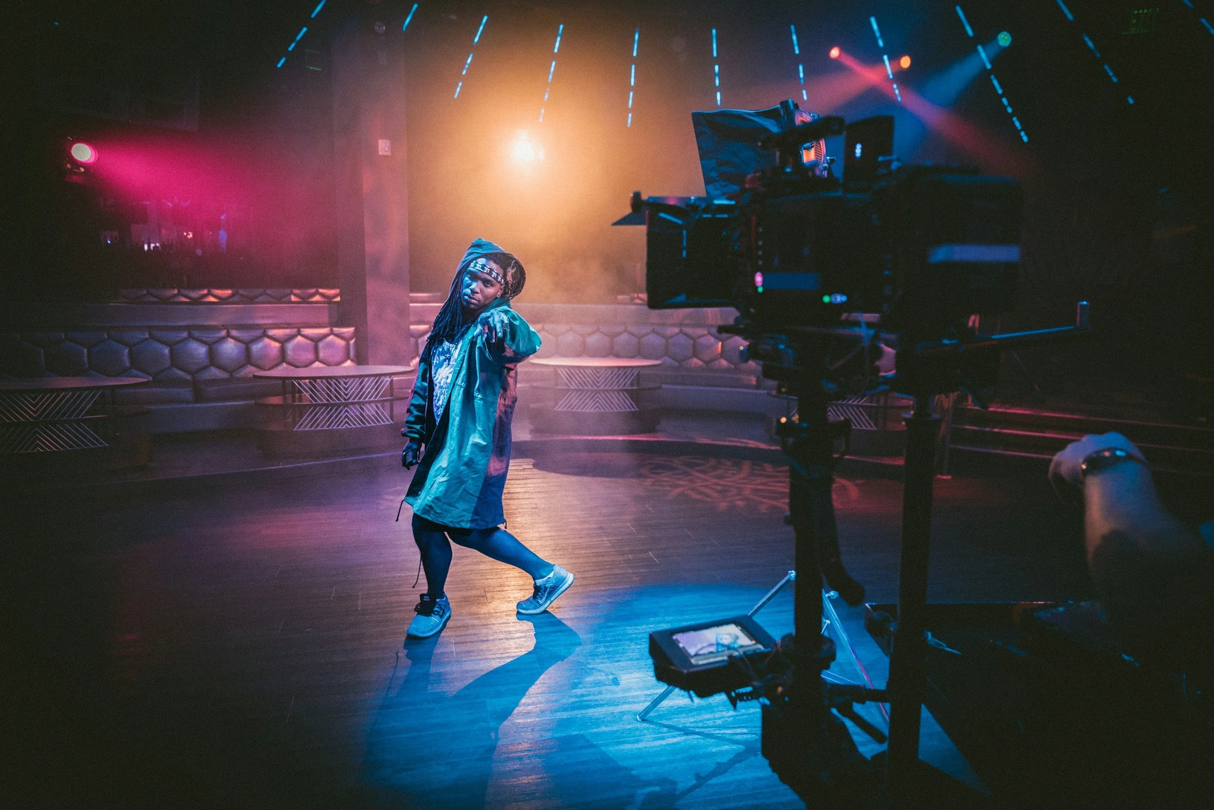 A person with dreadlocks wearing a blue jacket and shorts, posing in a dimly lit dance or performance venue with stage lighting and equipment visible.