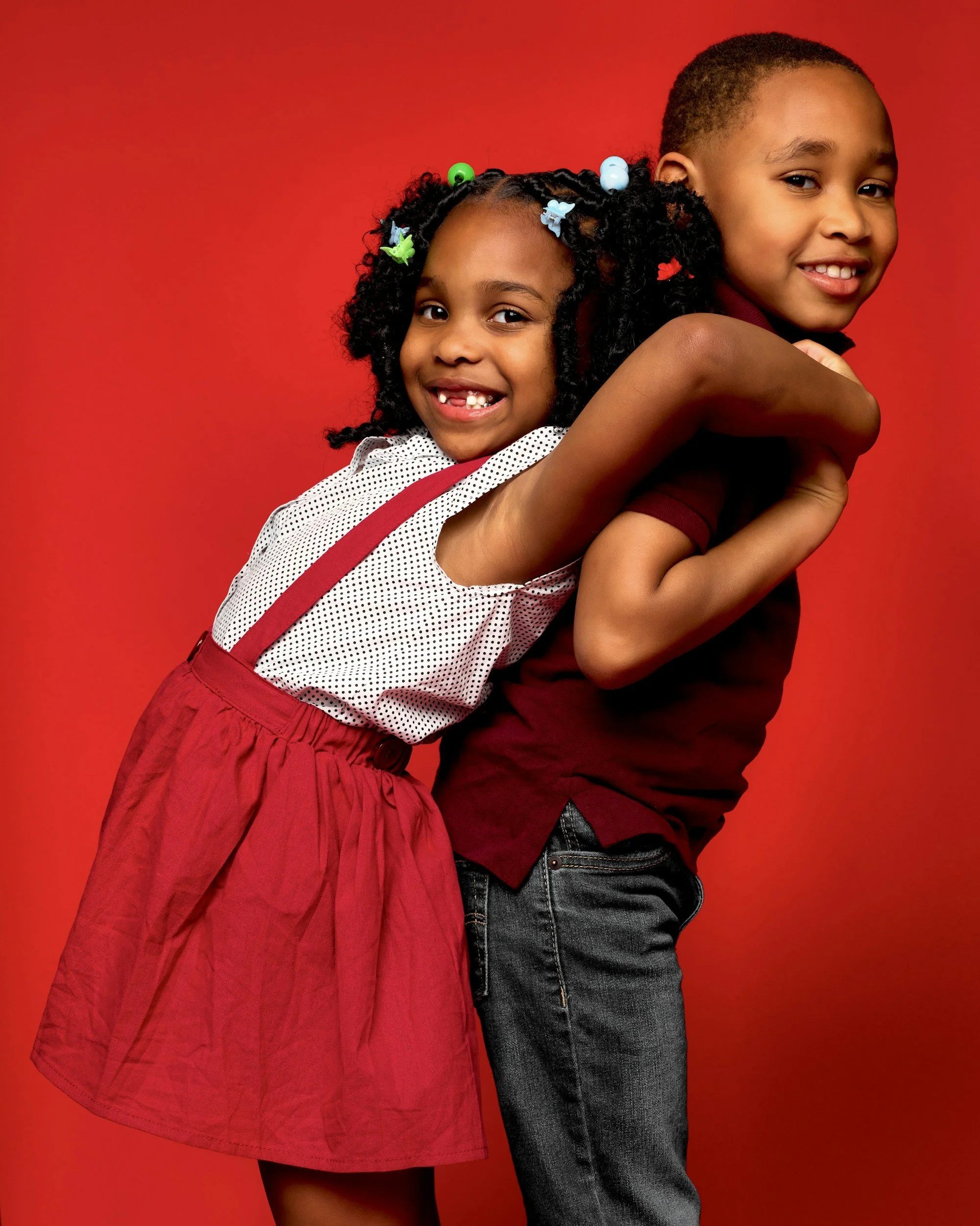 Smiling young girl with curly hair decorated with colorful beads, being embraced by a smiling teenage boy, against a red background.