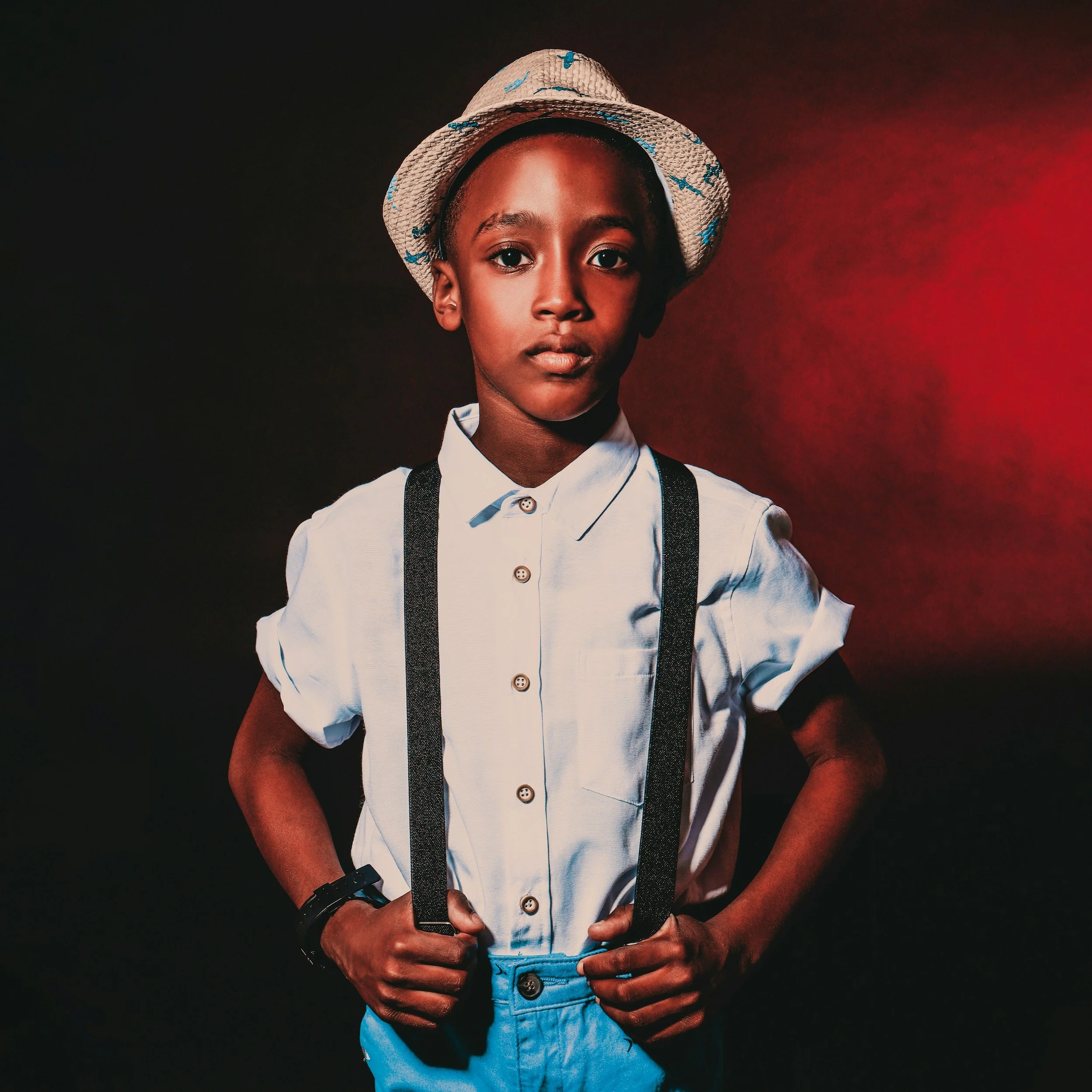 A young person wearing a straw hat with blue accents, a white button-up shirt, suspenders, and a black watch, holding a hat in front of a dark background with red lighting.
