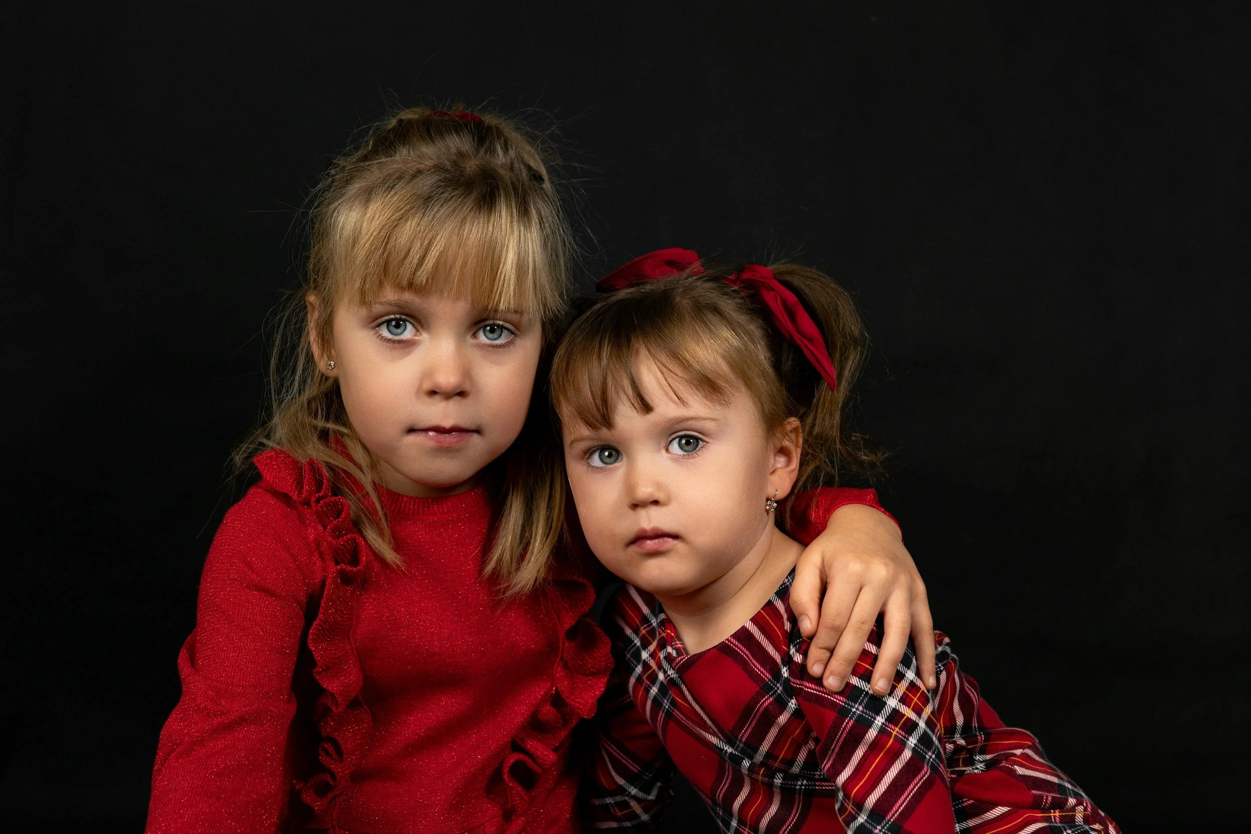 Two young girls with blonde hair and blue eyes, dressed in red and plaid dresses, sitting together against a black background. The older girl has her arm around the younger girl, and both are looking directly at the camera.