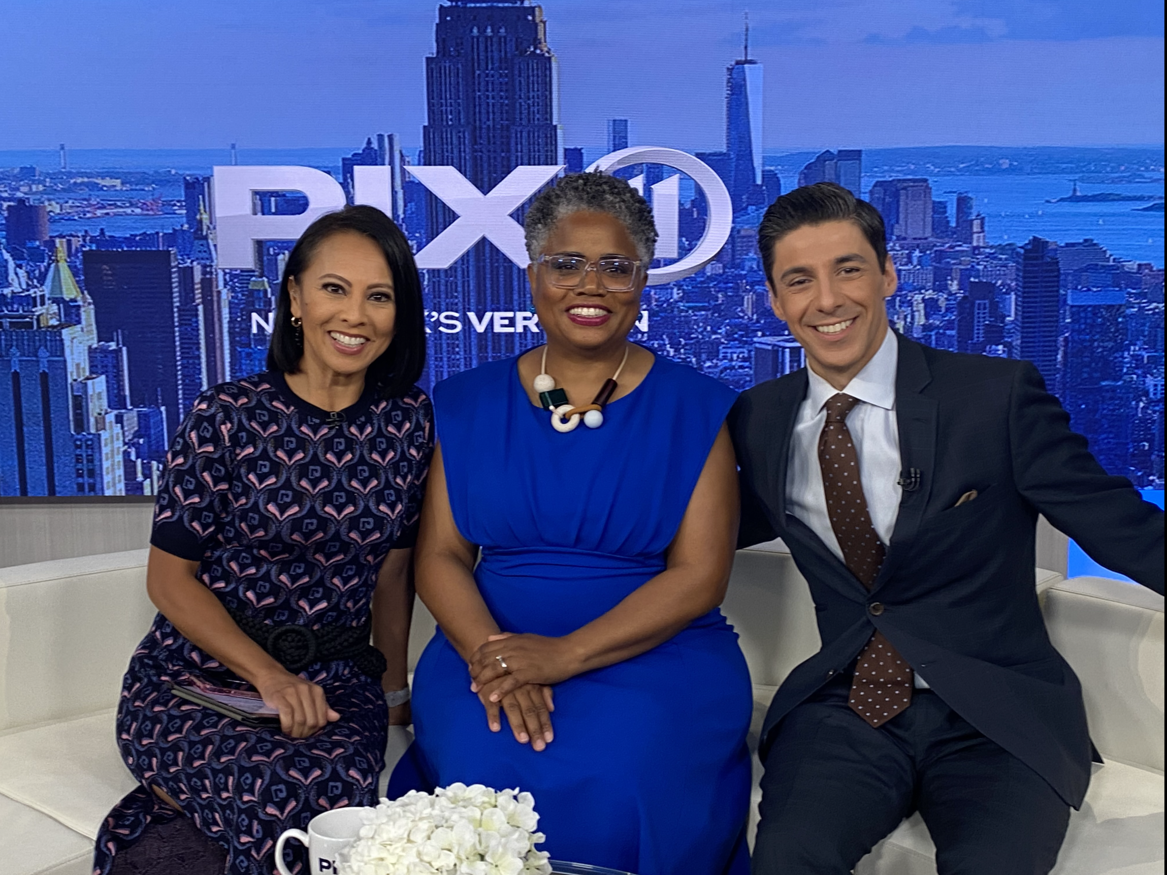 Three news anchors sitting in a studio with a cityscape background featuring skyscrapers and a river. They are smiling, with a table of white flowers and mugs in front of them.