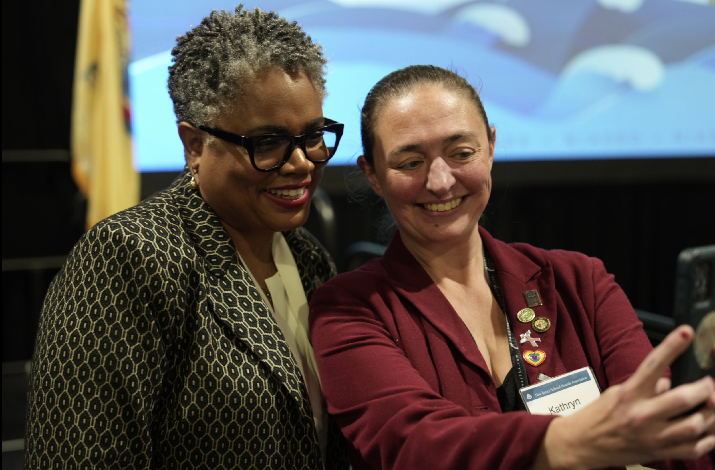 Two women are smiling and taking a selfie together at an event. The woman on the left has short, curly gray hair, wears glasses and a patterned blazer. The woman on the right has dark hair pulled back, is wearing a maroon blazer with pins, and has a 