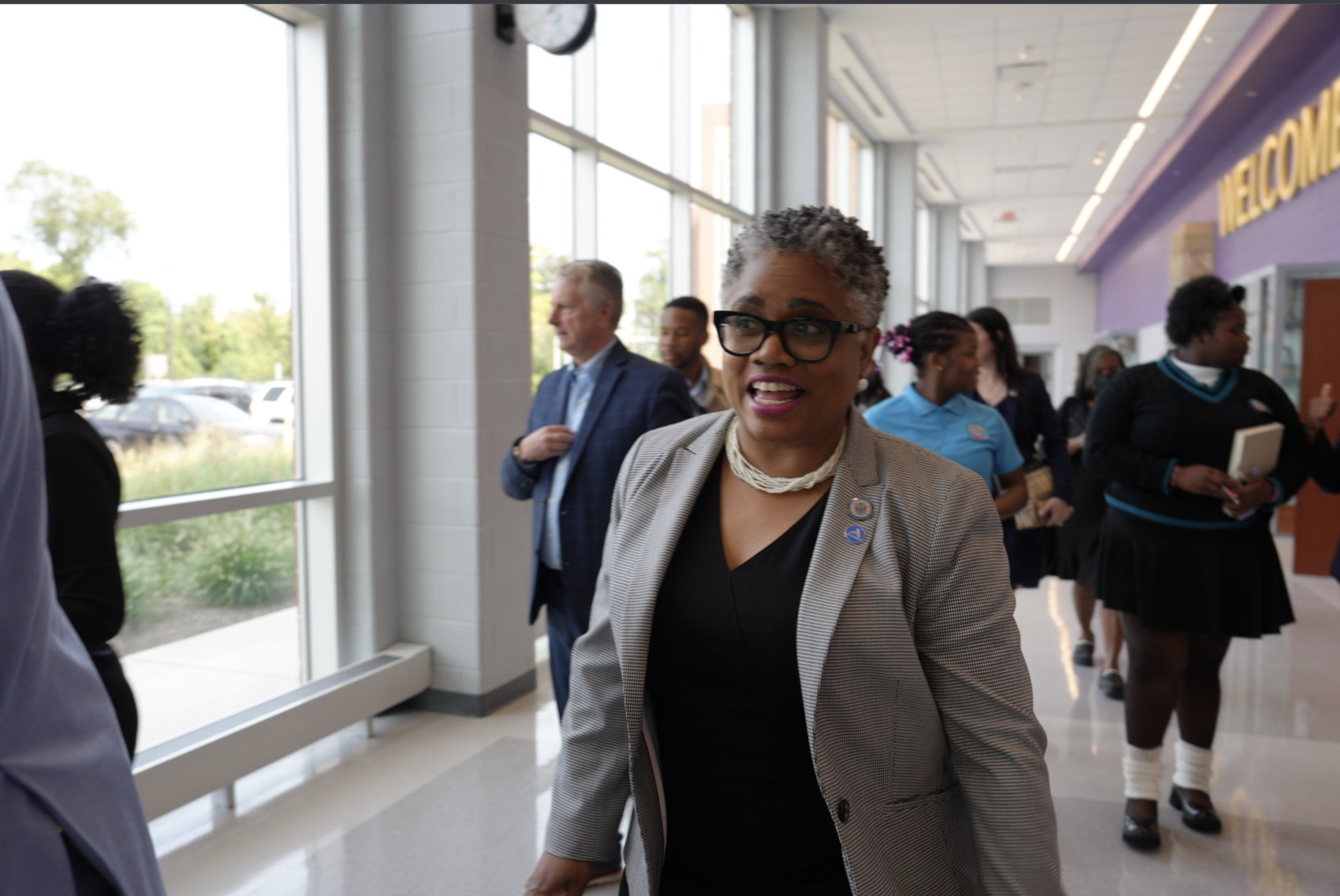 A woman with short curly gray hair, black glasses, and a beige blazer walking through a modern indoor space with large windows and other people in the background.