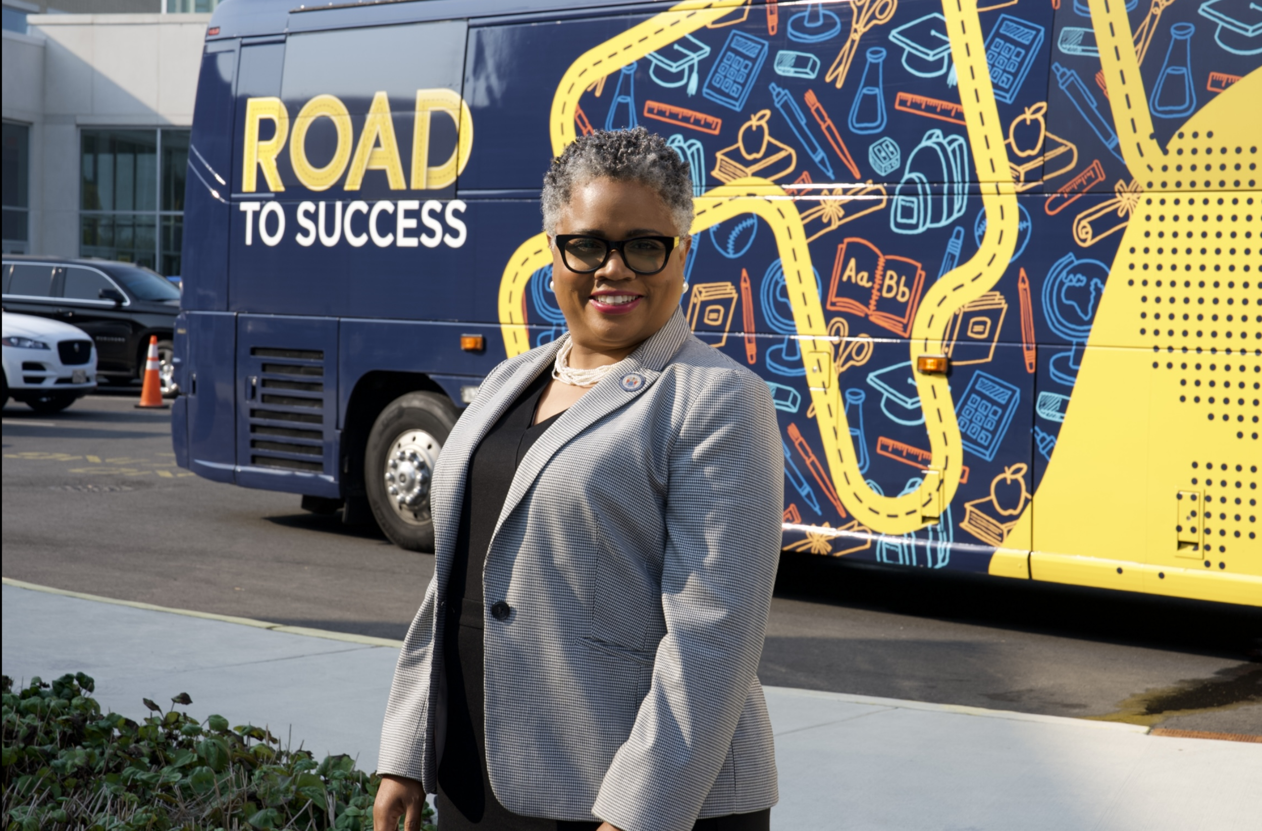 Woman with short curly gray hair and glasses smiling in front of a mural and a bus that says 'Road to Success'