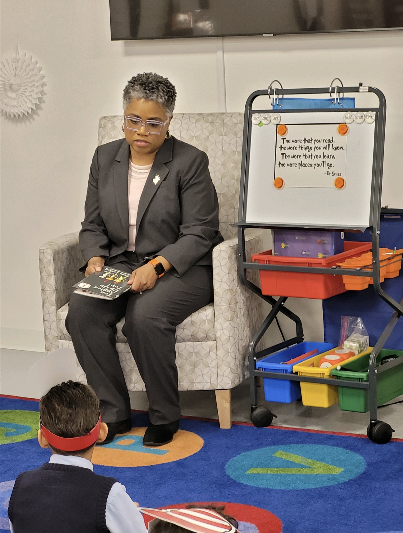 A woman with short, curly gray hair and glasses, dressed in a gray suit, sitting on a patterned armchair, reading a children's book during a classroom activity. In front of her, children sit on a colorful rug, one with a red headband, engaged in a st
