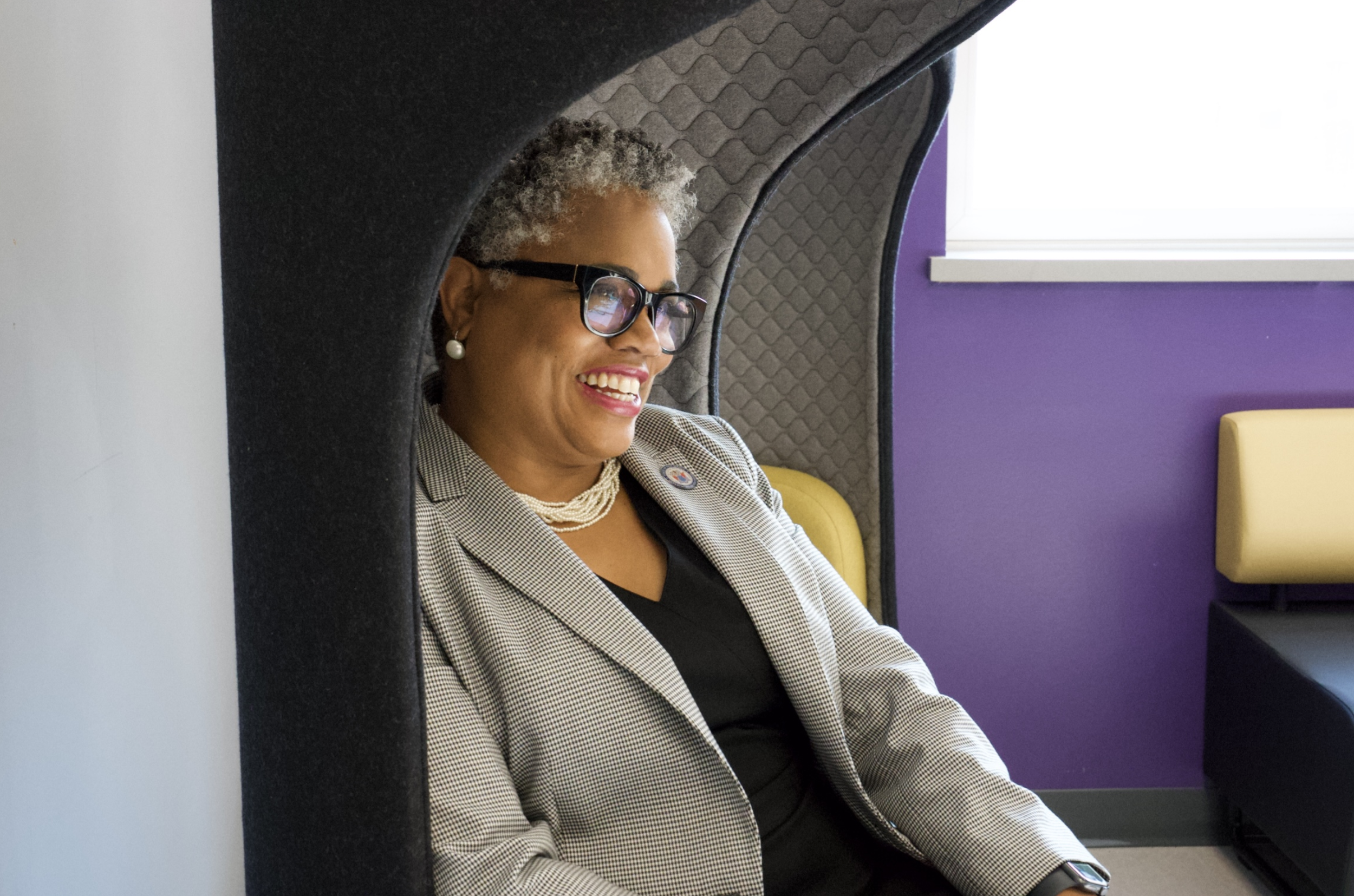A woman with curly gray hair, wearing glasses, pearl earrings, a checkered blazer, and a black top, is sitting on a yellow chair, smiling. She is seen through a cozy, enclosed space with a curved, padded fabric structure around her, with a purple wall behind her.