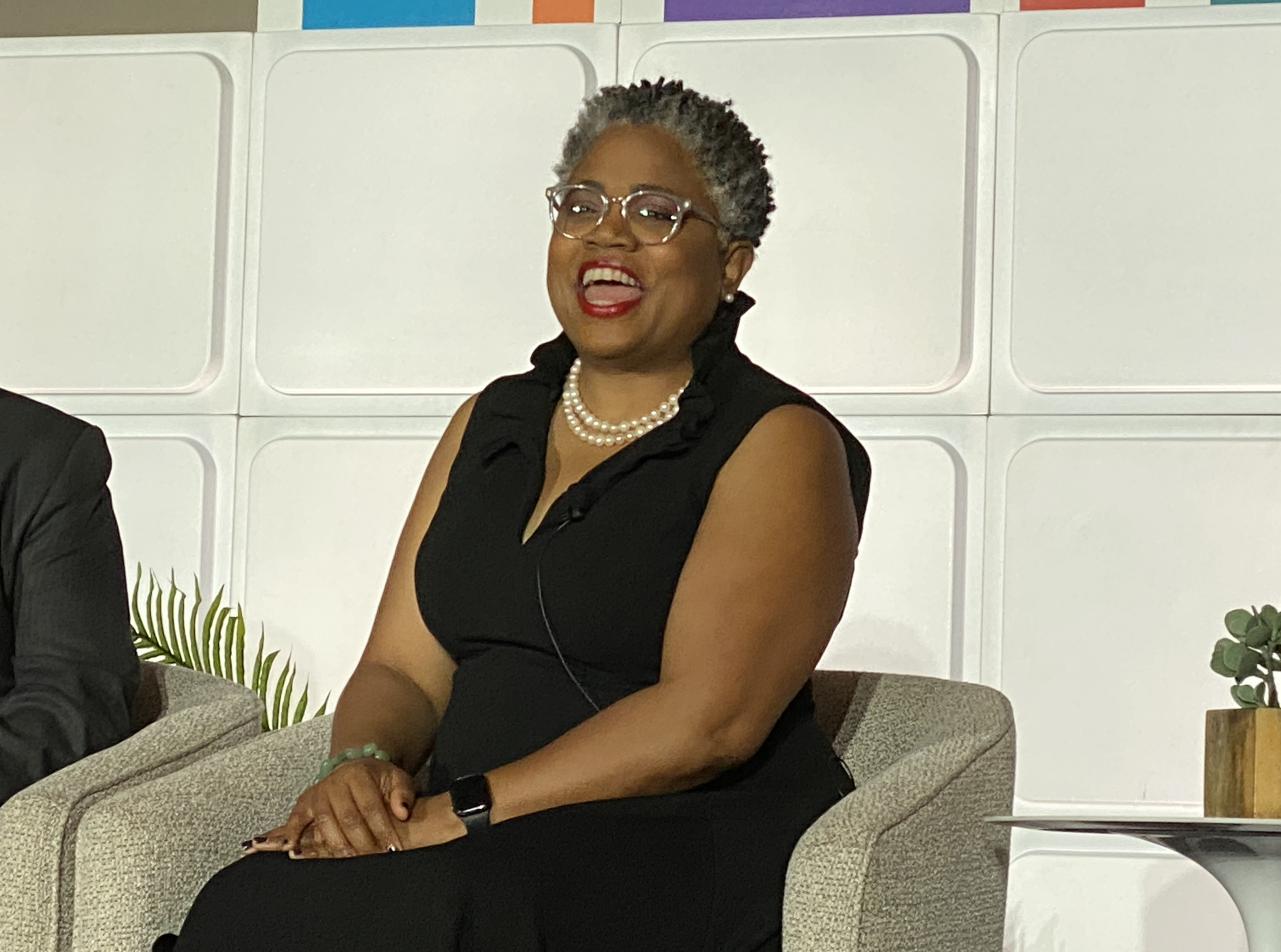 A woman with short gray curly hair, glasses, and a pearl necklace is sitting on a couch. She is smiling and wearing a sleeveless black top. The background features a white wall with colorful accents.