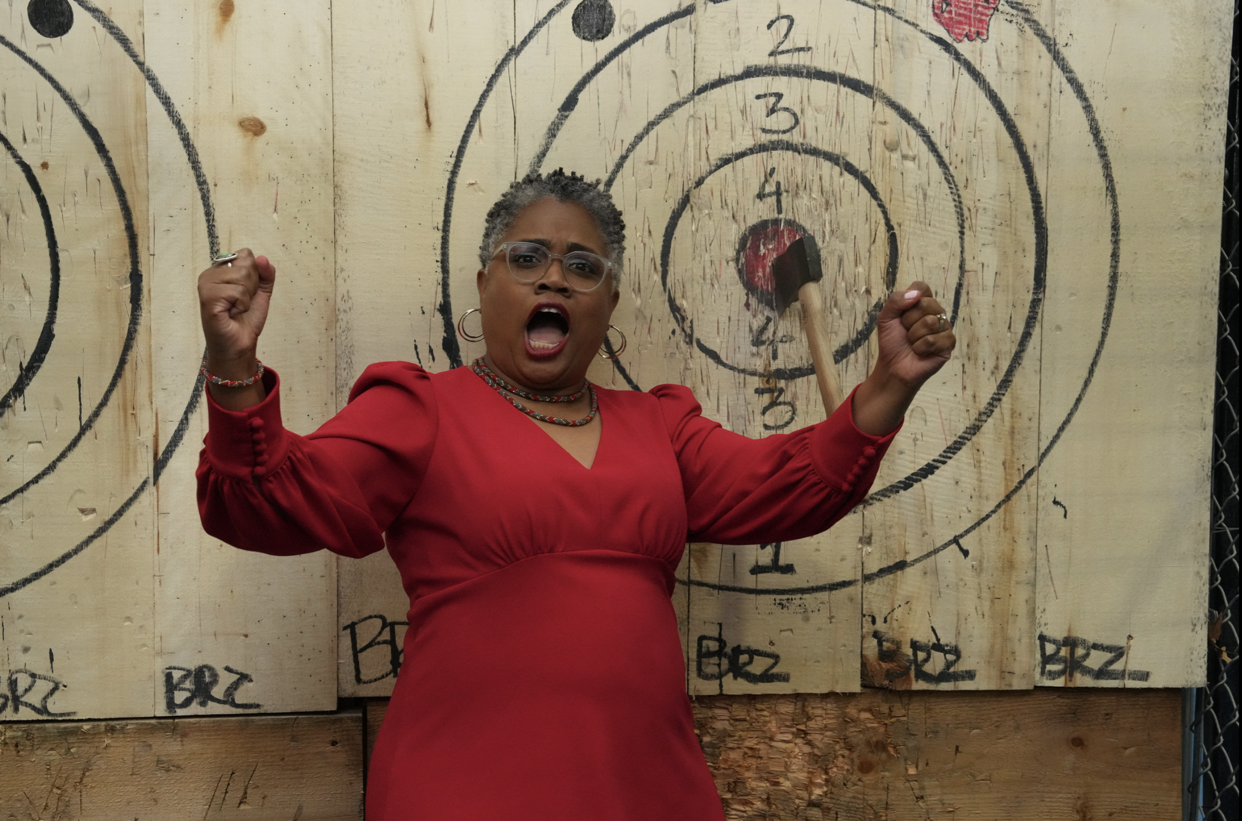 An older woman wearing glasses and a red dress is standing in front of a wooden target board with black concentric circles and numbers. She has her fists raised in a victorious pose, mouth open as if cheering.
