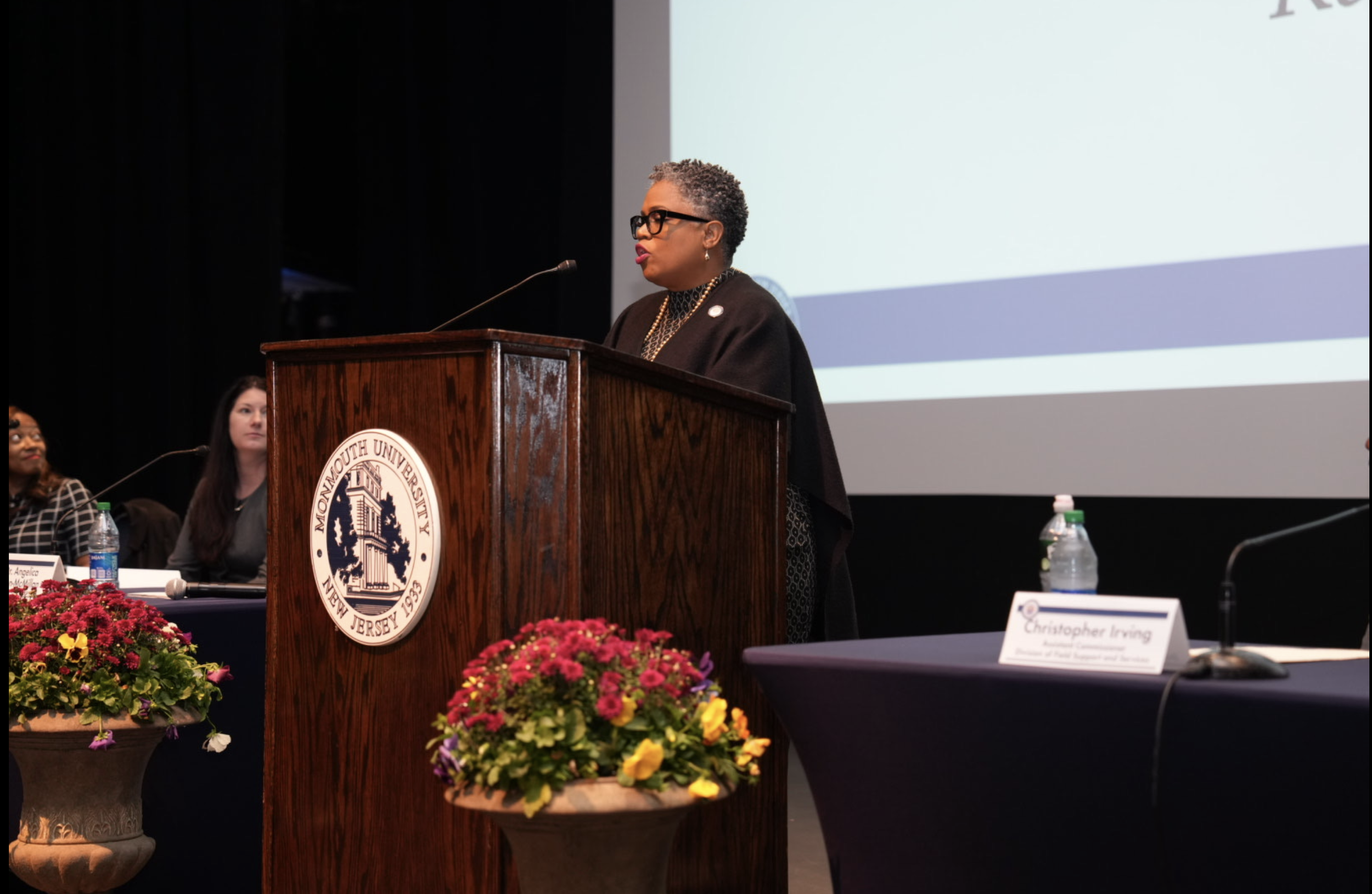 A woman with short gray hair and glasses speaking at a podium with the Monmouth University emblem, with women and floral arrangements in the background.