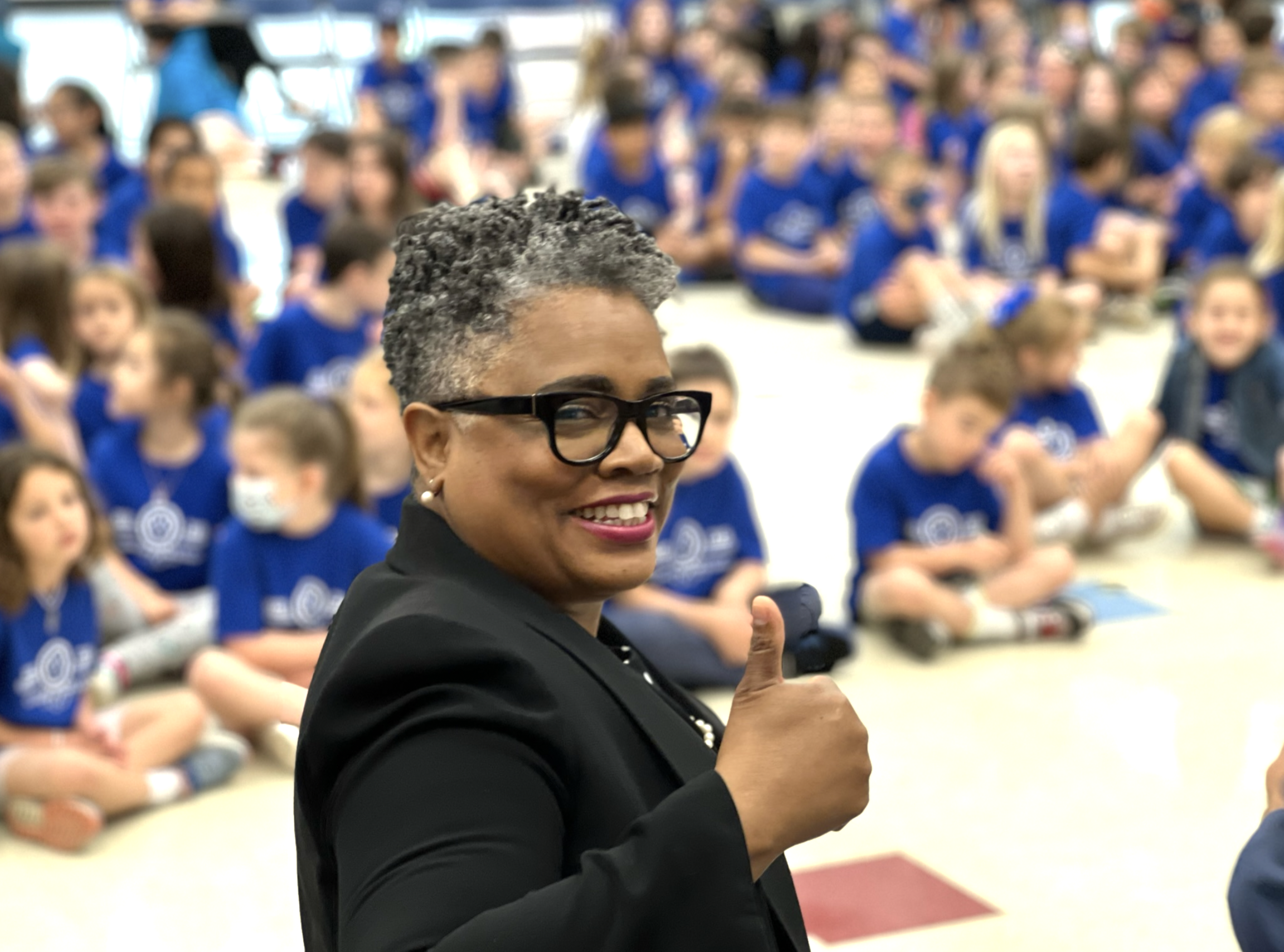 A woman wearing glasses and a black blazer giving a thumbs up in front of a group of children in blue shirts seated in a classroom or auditorium.