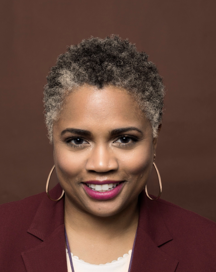 Professional woman with short, curly salt-and-pepper hair, wearing a burgundy blazer and hoop earrings, smiling against a brown background.