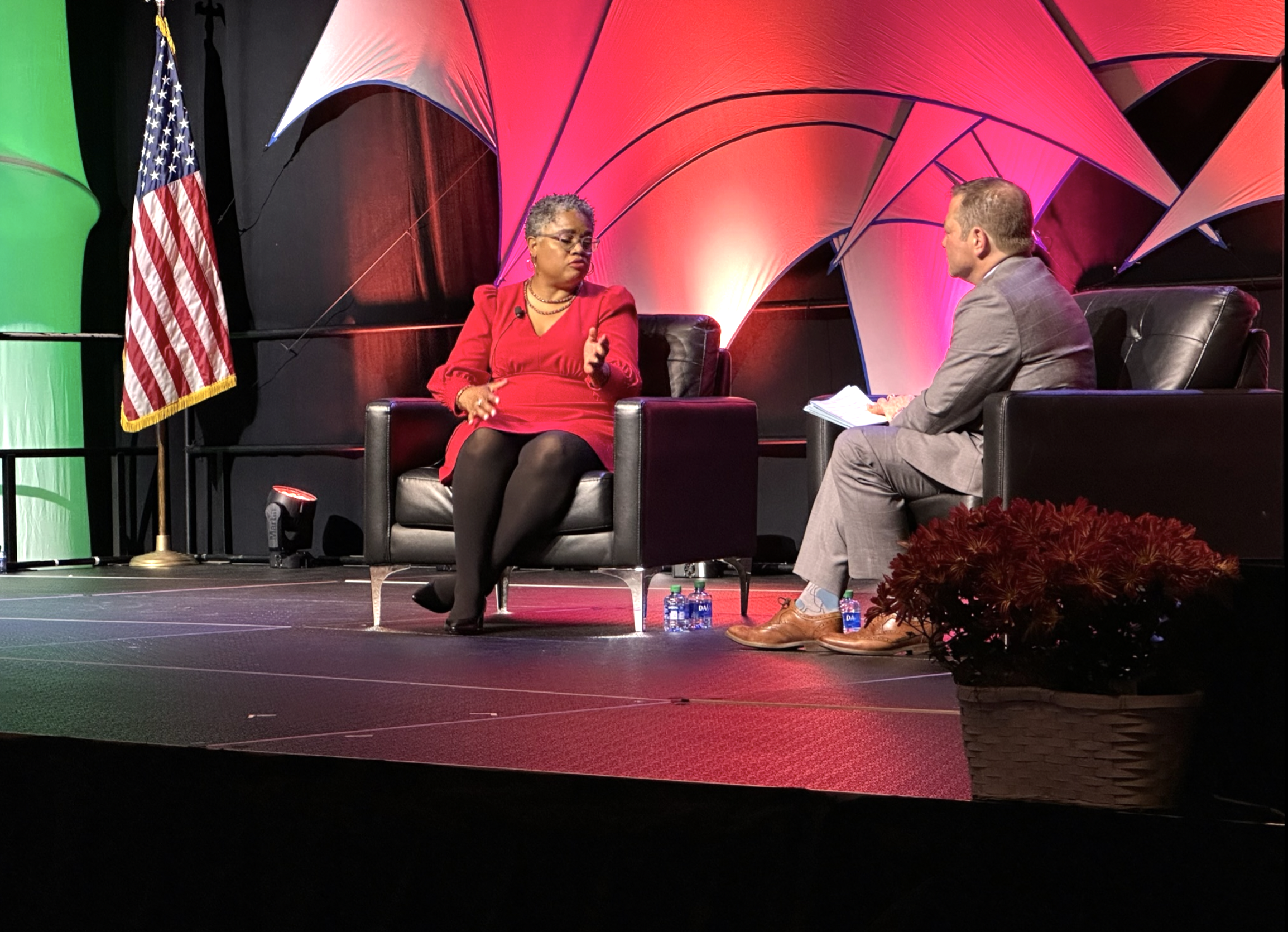 A woman in a red dress and glasses sitting on stage, talking to a man in a gray suit holding papers, with an American flag and a colorful abstract backdrop behind them.