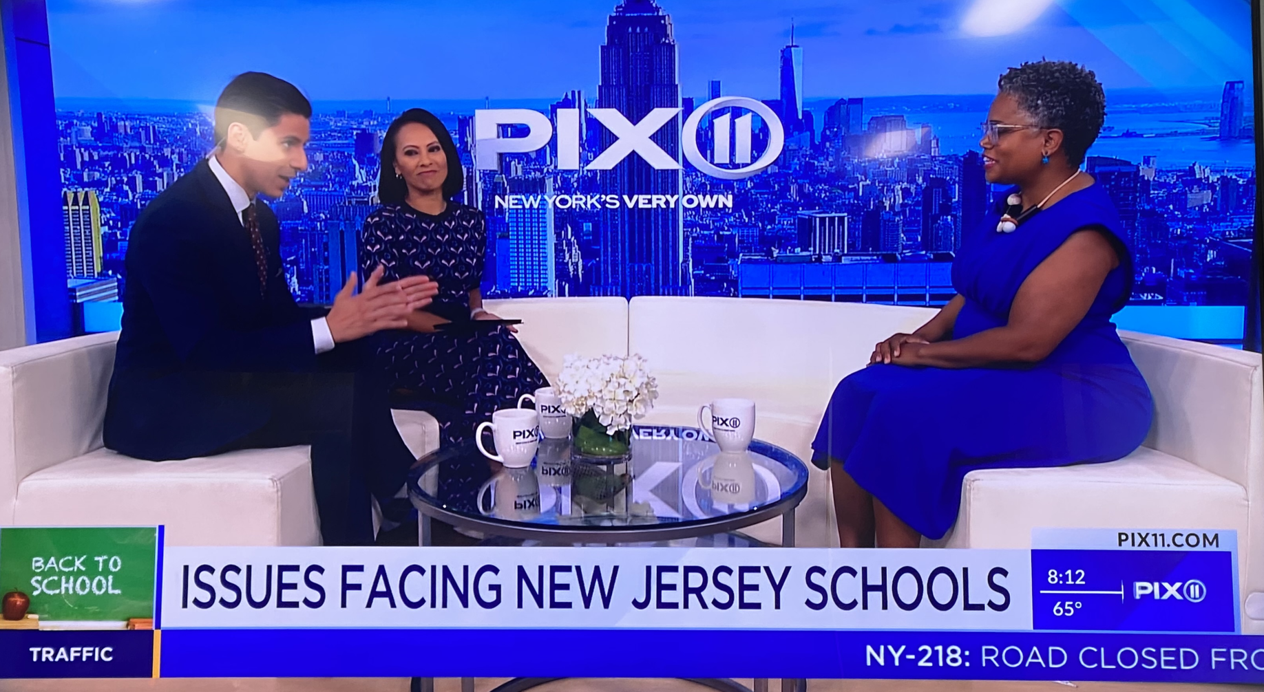 Three people sitting on a white studio couch engaged in conversation. The show logo 'PIX11' is visible on a large screen behind them, showing New York City skyline. A small round glass table with white flowers and PIX11 mugs is in front of them. A bo