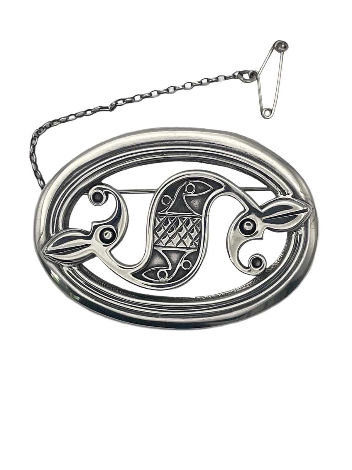 A round silver decorative piece with a chain attached at the top. The design features a central element with a diamond pattern, flanked by two stylized birds facing opposite directions, with detailed features.