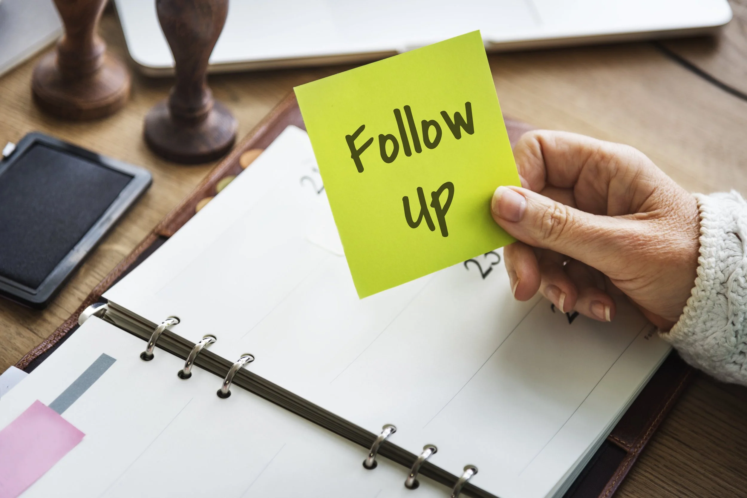 A person's hand holding a yellow sticky note that says 'Follow up' in black letters. In the background, there are wooden clock figurines, a smartphone, and a planner on a wooden desk.