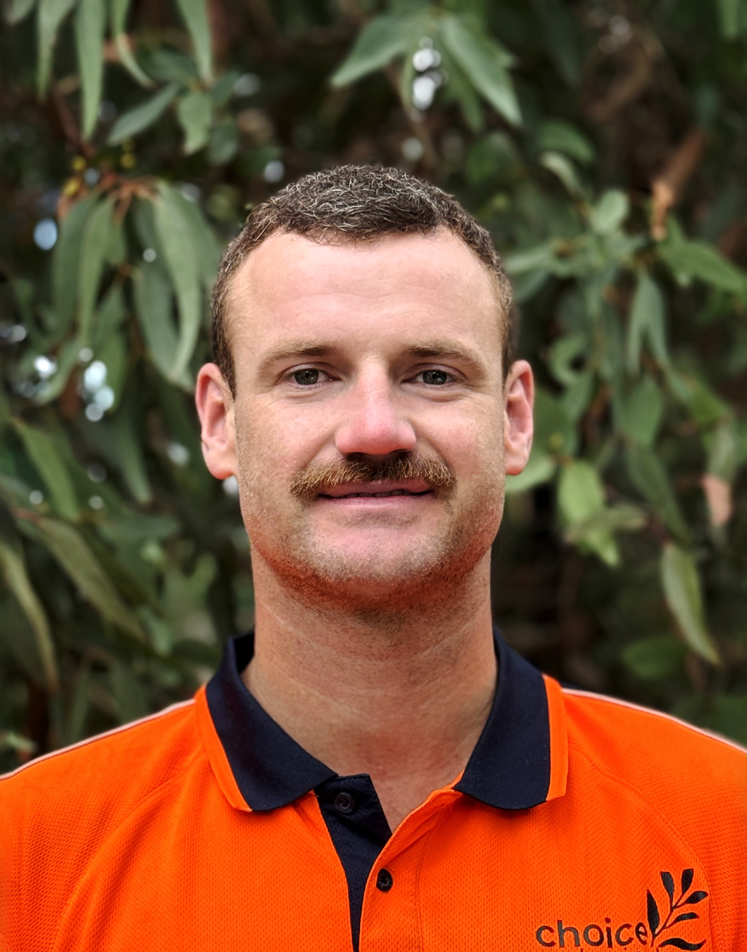 Evan Gwilym: a man with short, brown hair and a moustache, wearing an orange shirt with black accents, standing outdoors with green foliage in the background.