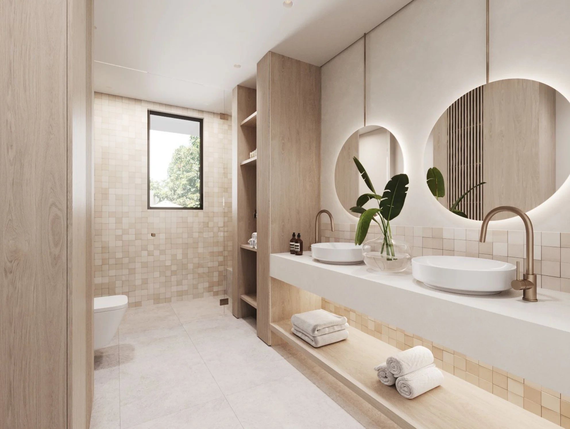 Modern bathroom featuring dual white vessel sinks with brass faucets, a large circular mirror with backlighting, a countertop with a plant and towels, and a walk-in shower with beige tiles and a window showing outdoor greenery.