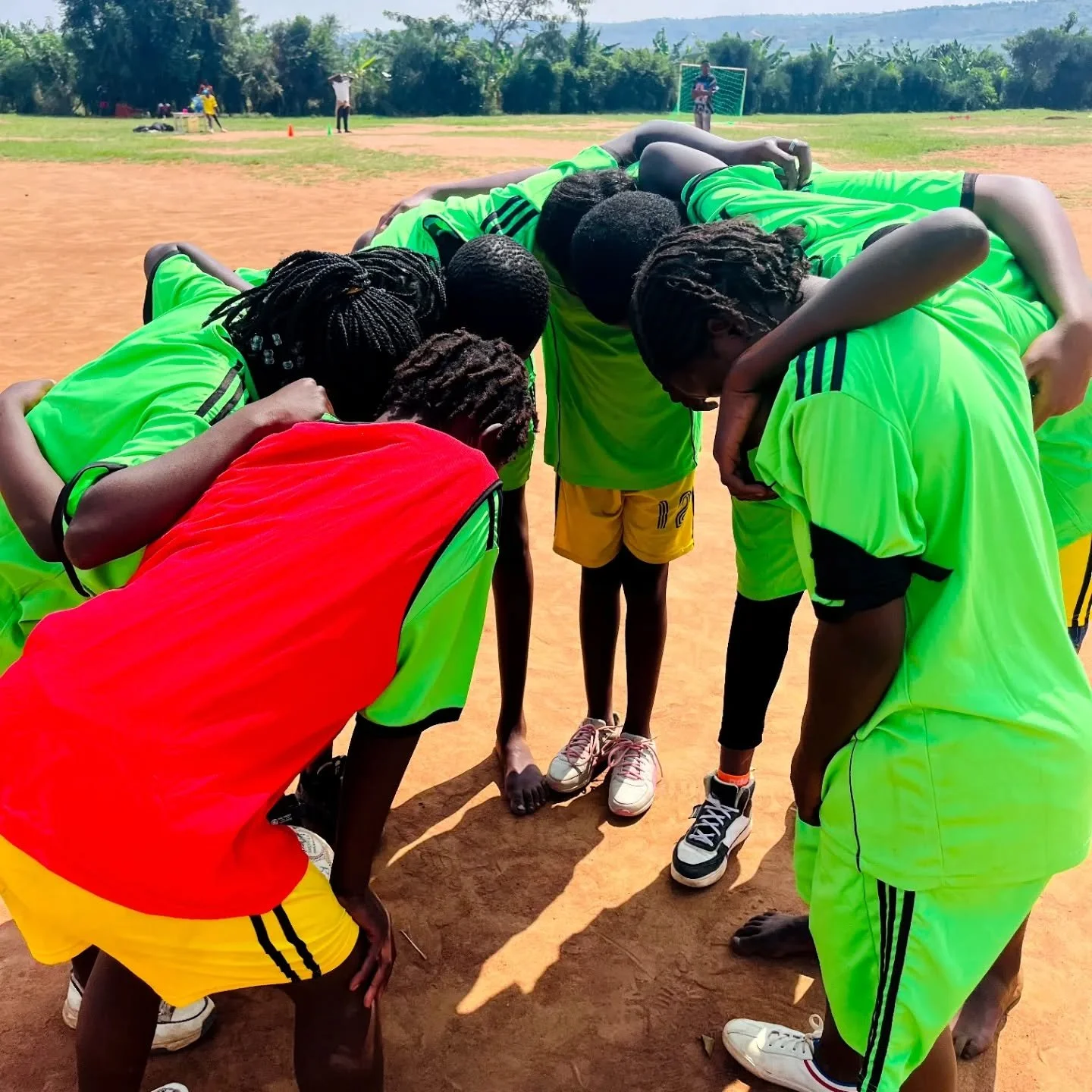 A group of young soccer players in bright green and red jerseys huddled together in a circle on a dirt field, preparing for a game.