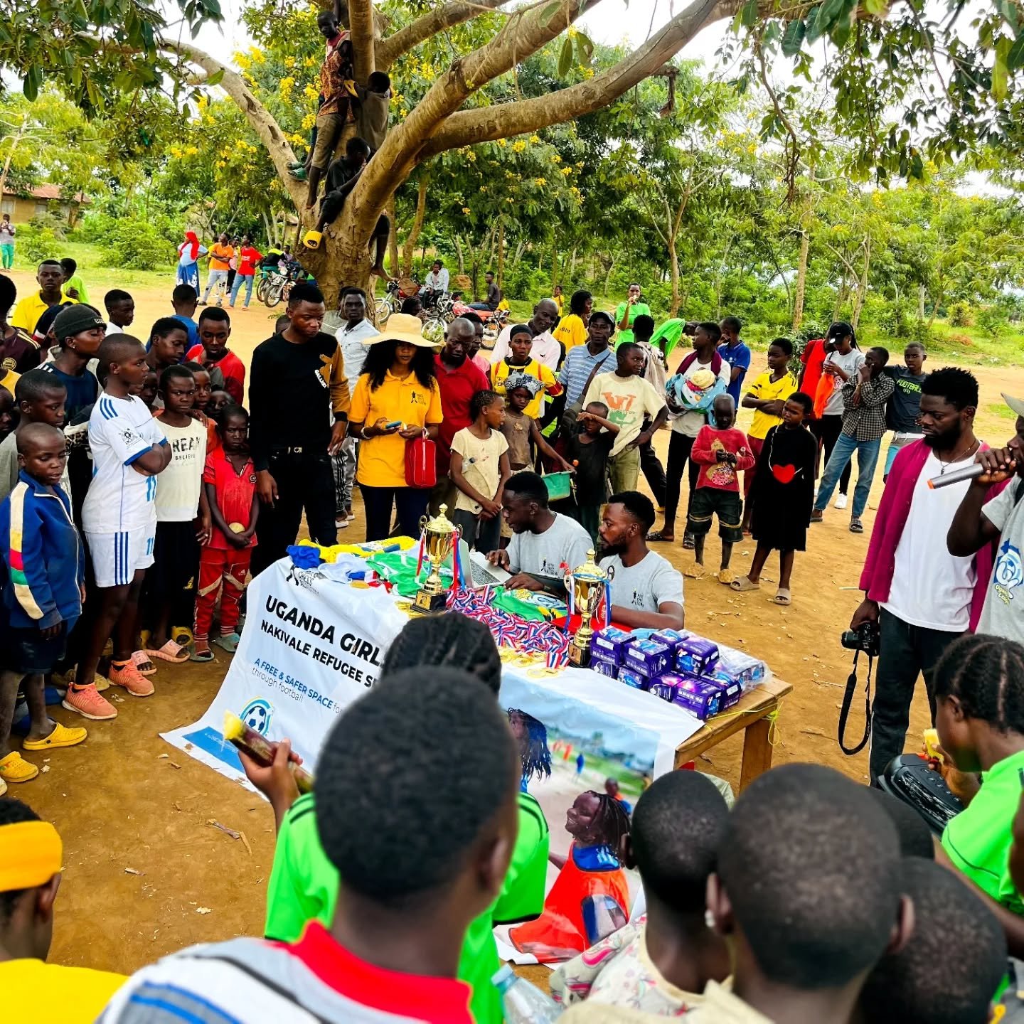 A community event outdoors with a crowd of people gathered around a table displaying trophies, medals, and sports equipment under a tree during the day.