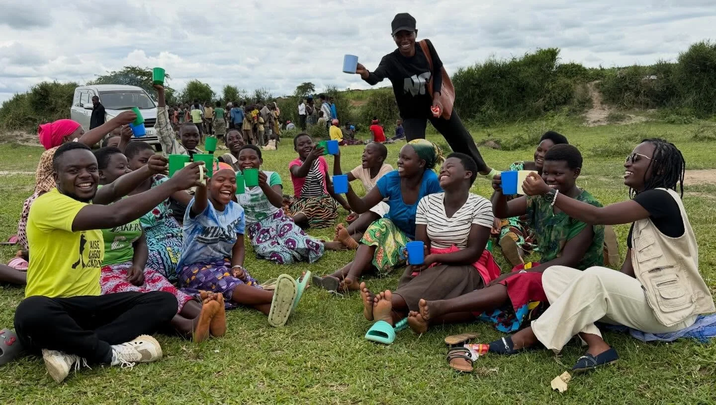 Group of people in colorful clothing sitting on grass, raising cups, celebrating outdoors, with a woman standing above them holding a cup, in a rural area with trees and a cloudy sky in the background.