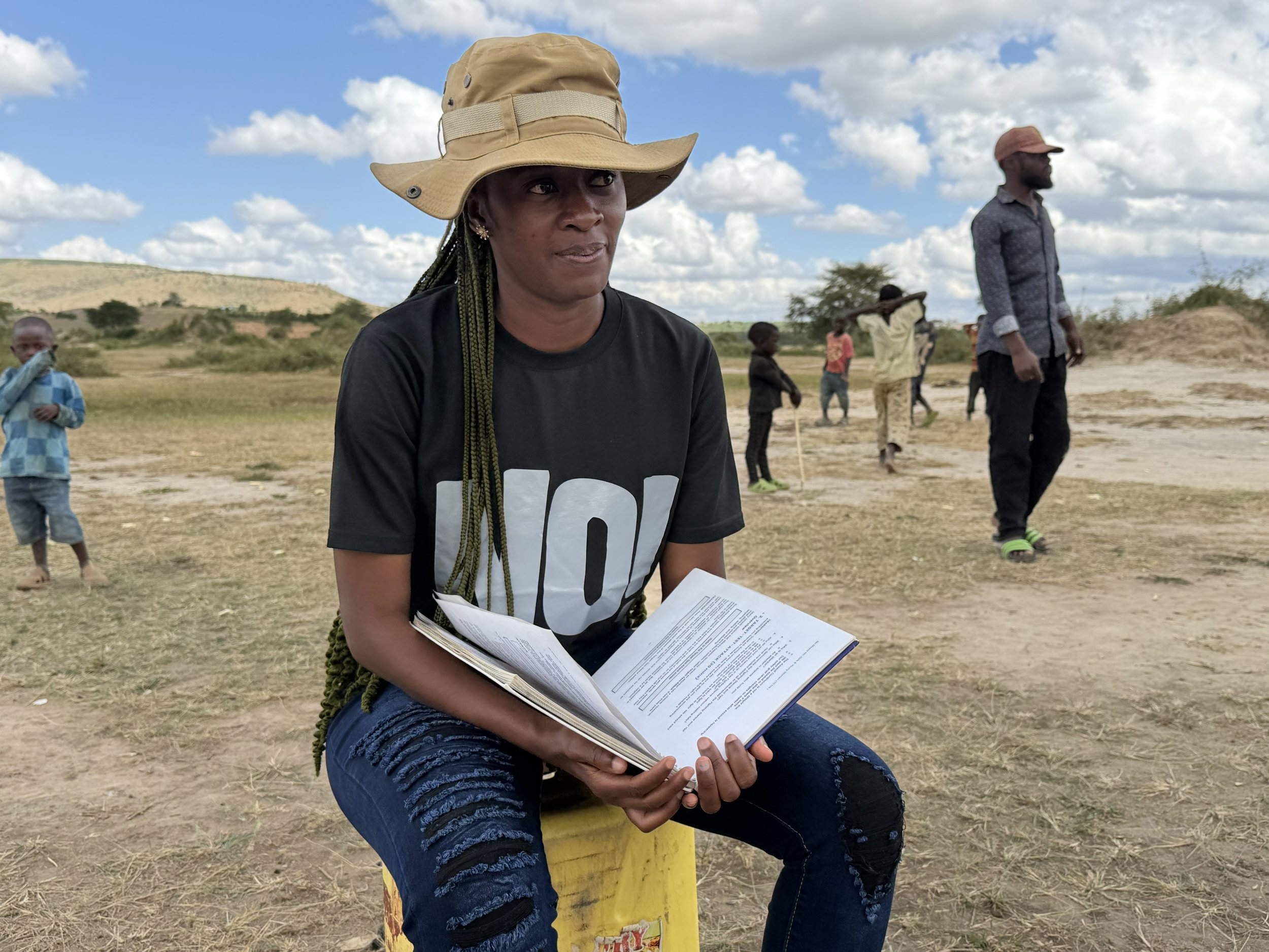 A woman wearing a tan wide-brimmed hat and black T-shirt with white lettering, sitting on a yellow bollard, holding papers, outdoors on a sunny day with partly cloudy sky. Several children and a man are in the background, walking or standing on a grassy landscape.